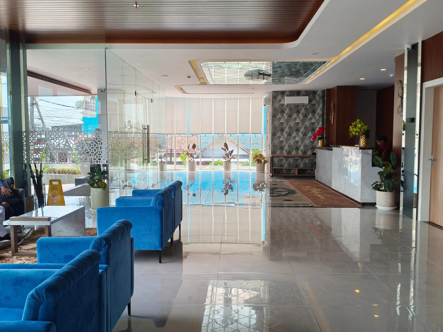 Hotel lobby with blue velvet chairs, white marble tables, potted plants, a reception desk with gold vases and red and green plants, and a swimming pool visible through large glass windows at the back.