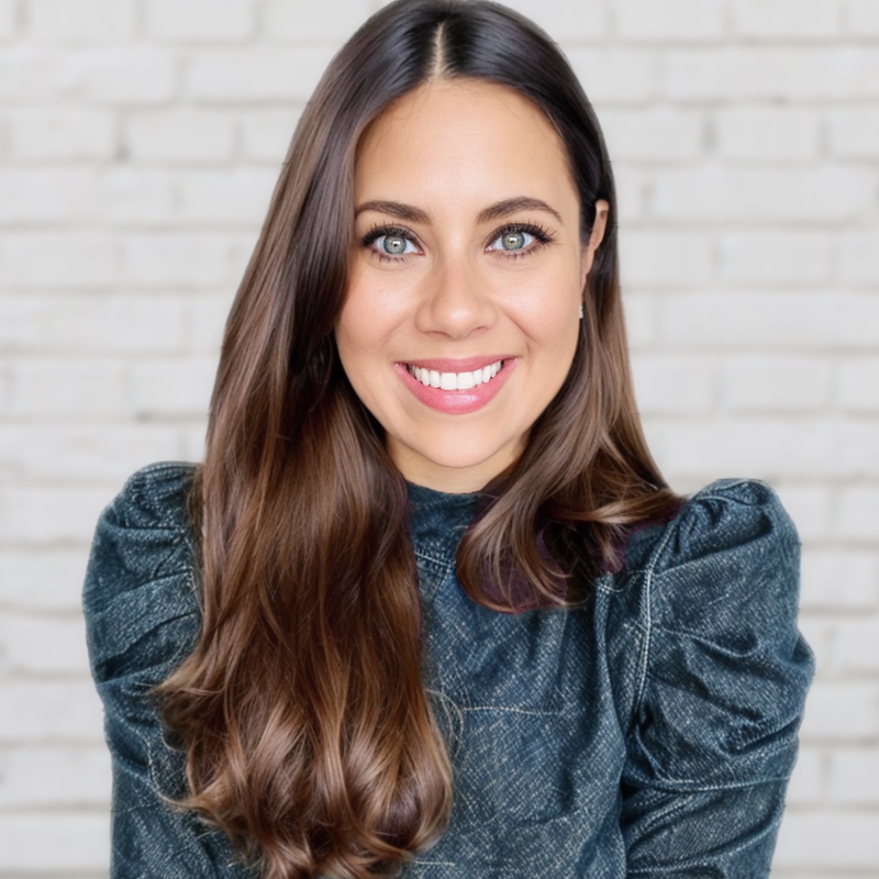 A woman with long brown hair, blue eyes, and a bright smile, wearing a blue denim top, standing in front of a white brick wall.