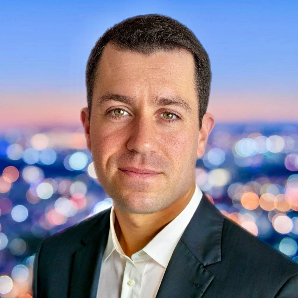 Headshot of a man in a suit with a city skyline background at dusk.