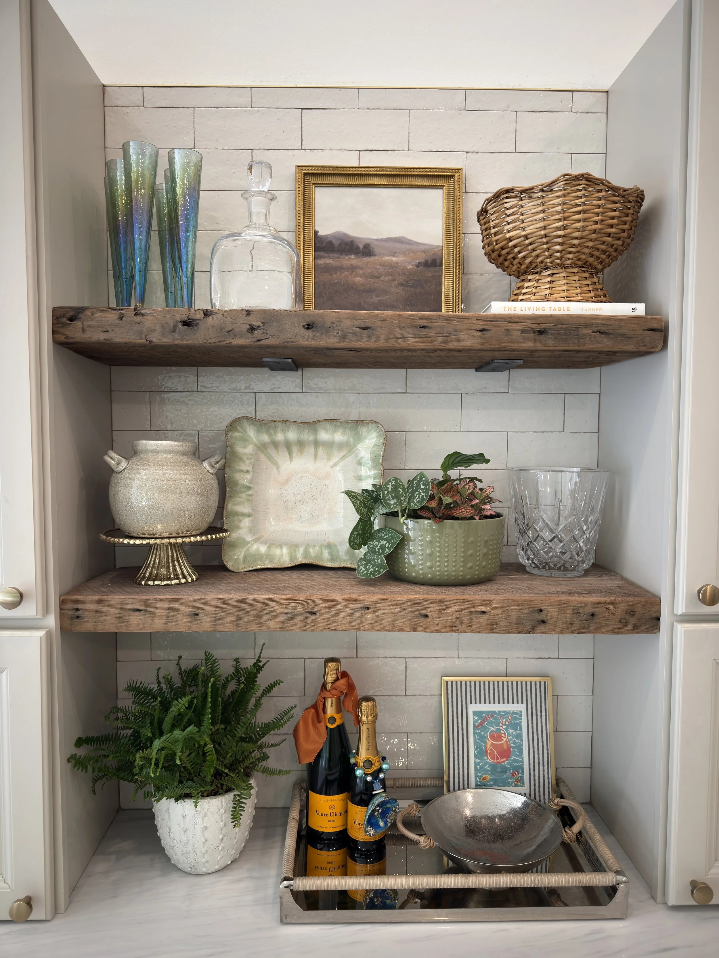 A decorative kitchen display shelf with various items including colorful glass vases, a glass decanter, a framed landscape painting, woven basket, books, potted plants, bottles of champagne, and a metal bowl, set against a white brick wall.