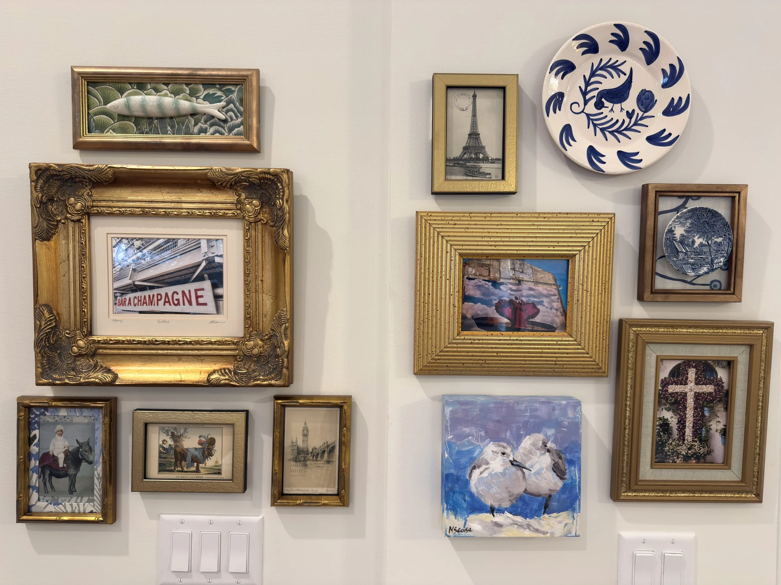 A gallery wall with various framed artworks, including a small ceramic fish sculpture on a green patterned background, a black-and-white photograph of the Eiffel Tower, a large white ceramic plate with blue bird and floral designs, a gold ornate fram