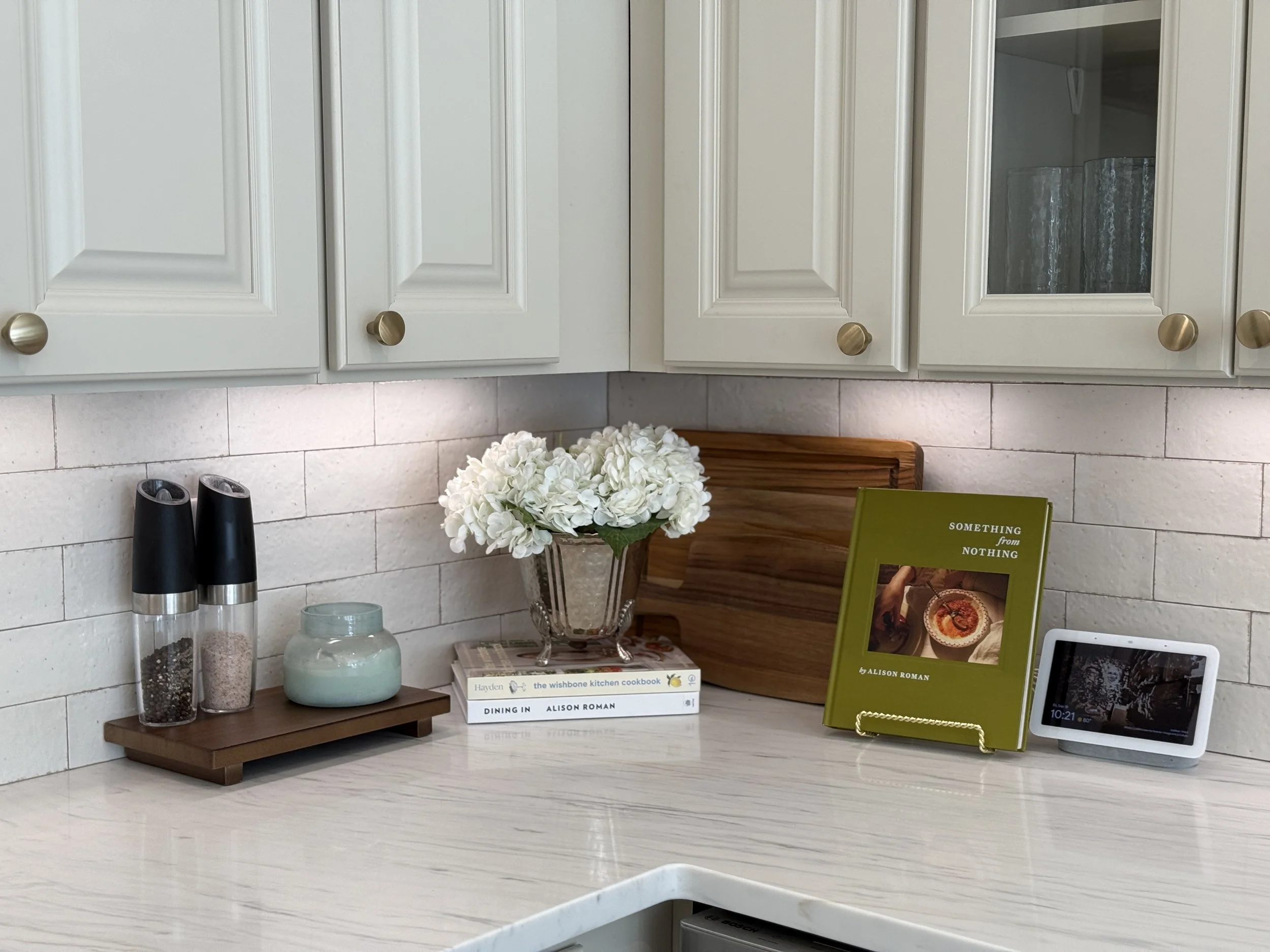 Kitchen shelf with white cabinets, a bouquet of white flowers, two black salt and pepper shakers, a light blue ceramic vase, a stack of cookbooks, a green book stand with a book titled 'Something from Nothing,' and a digital device displaying the tim