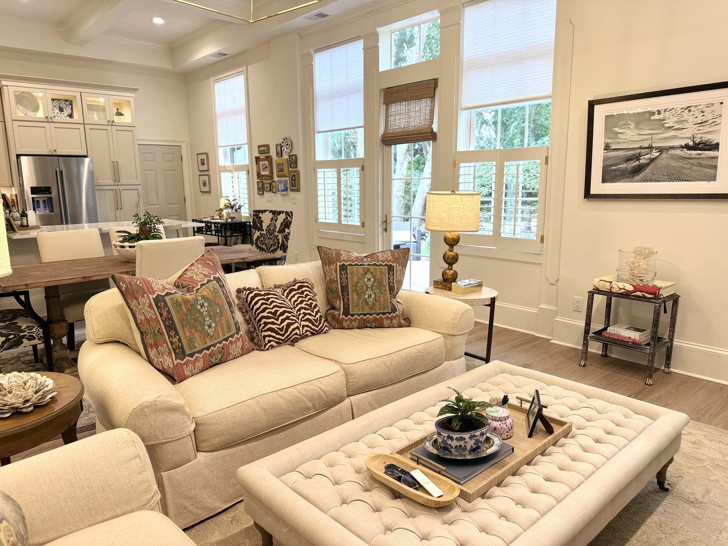 Living room with beige sofa, colorful patterned cushions, coffee table with plants and remote controls, and large windows with white blinds and bamboo shades.