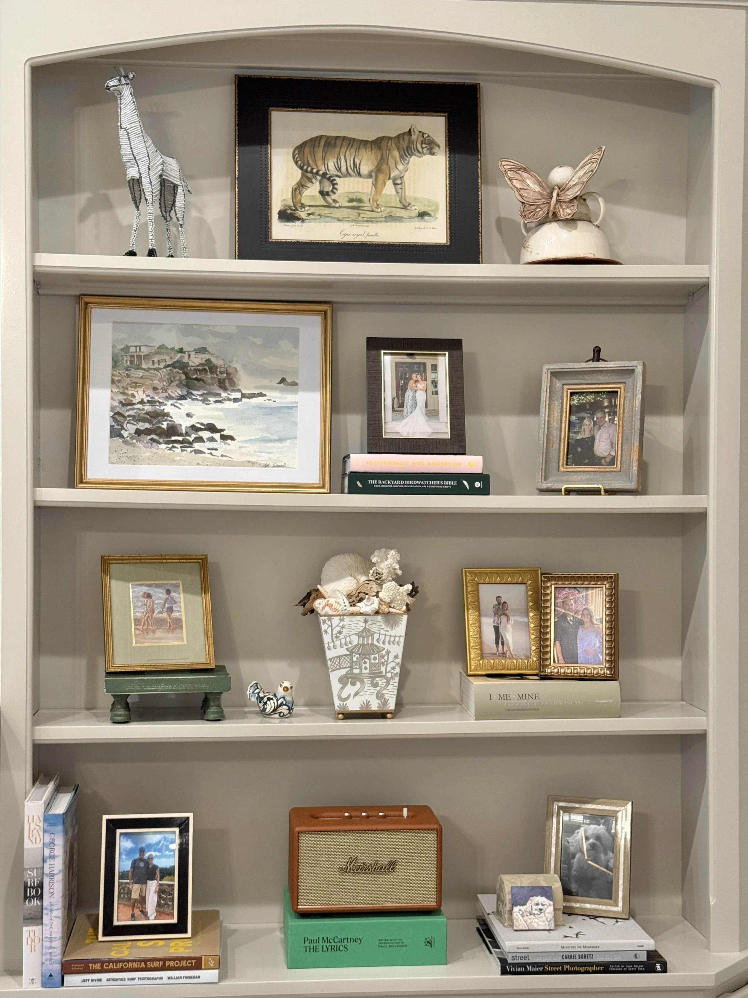 White bookshelf with various framed photos, artwork, books, and decorative items including a giraffe figurine, a tiger illustration, a butterfly-shaped ornament, a small animal figurine, a Marshall speaker, and a lucky cat figurine.