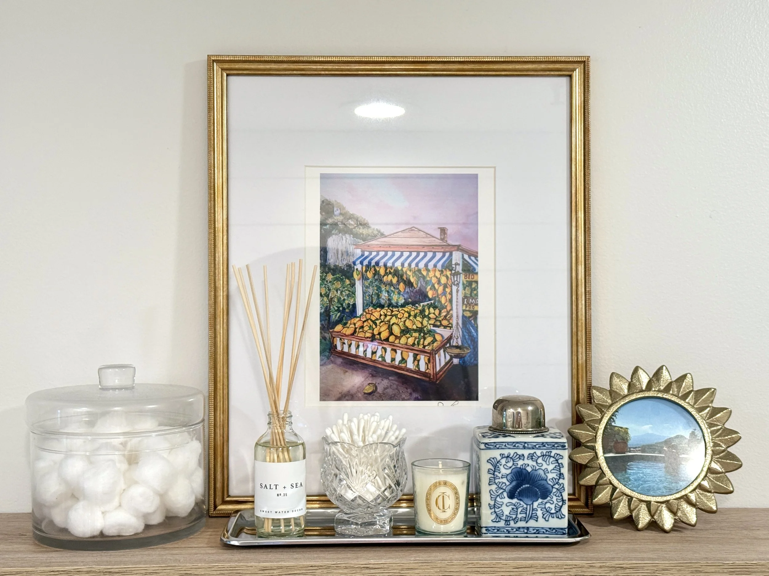 A decorative set of items arranged on a wooden surface, including a glass jar labeled 'Salt + Sea,' a glass bowl, candles, a framed picture of a scenic landscape, and a shiny golden decorative object.