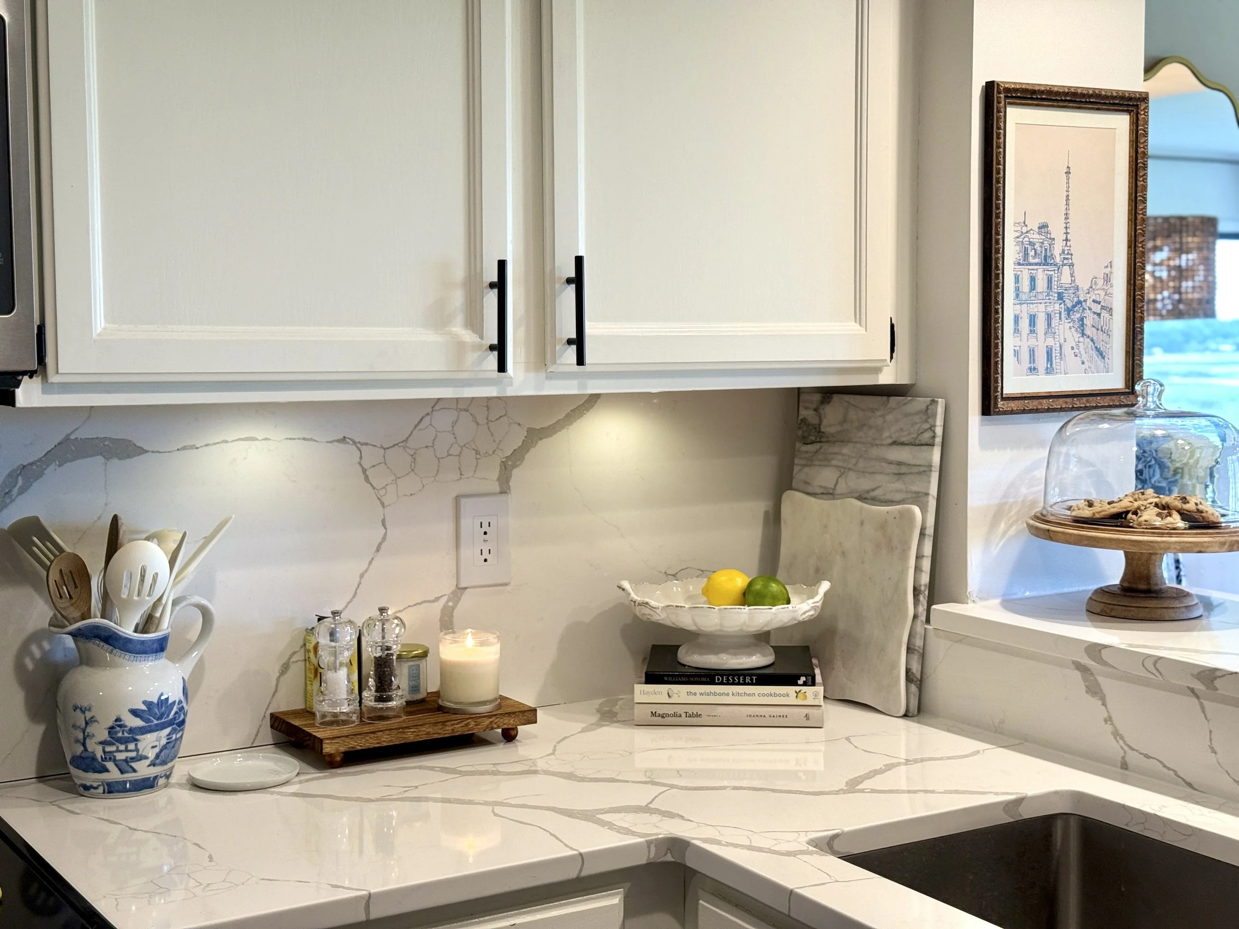A kitchen counter with a blue and white ceramic vase holding utensils, three small salt and pepper shakers, a lit candle, a stack of books, and a cake stand with cookies. Above, there are white cabinets and a framed picture of the Eiffel Tower.