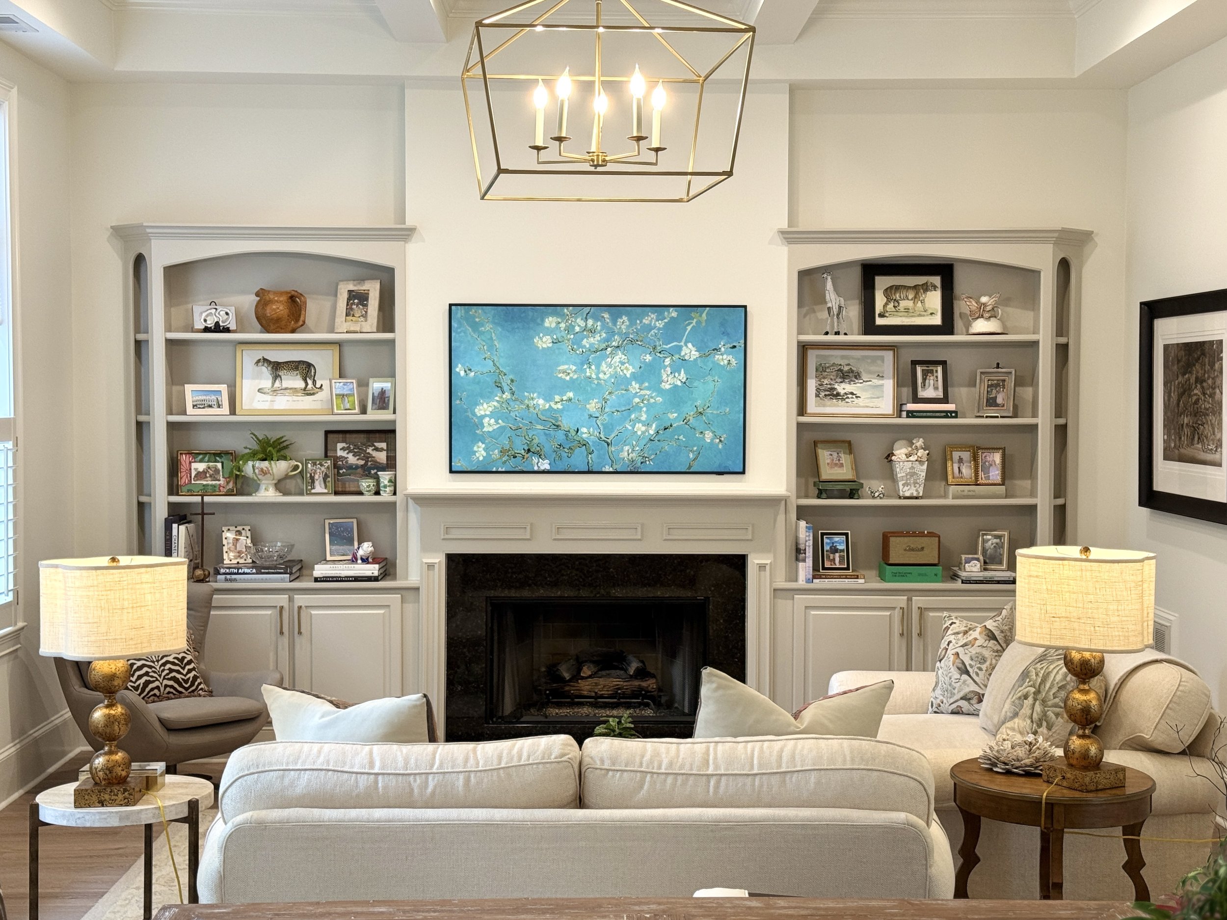 Living room with white sofa, armchair, patterned cushioned pillow, and side table with lamp. White built-in shelves with framed photos, artwork, and decorative objects. Large blue artwork of cherry blossoms above fireplace. Gold geometric chandelier 