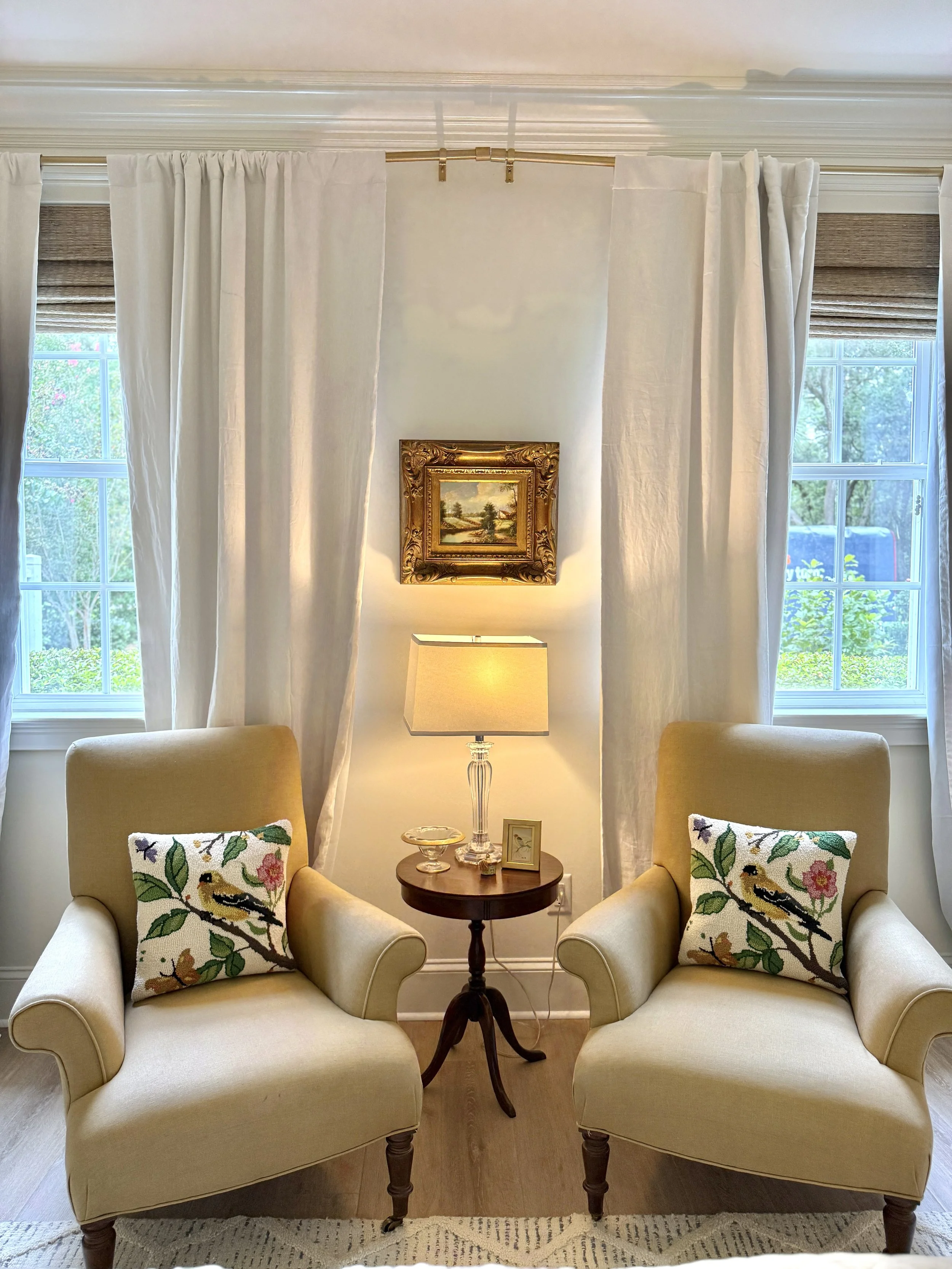 A cozy living room corner with two cream-colored armchairs, floral embroidered pillows, a small round wooden side table with a glass bowl, a picture frame, and a table lamp. Behind the armchairs are two large windows with white curtains and woven sha