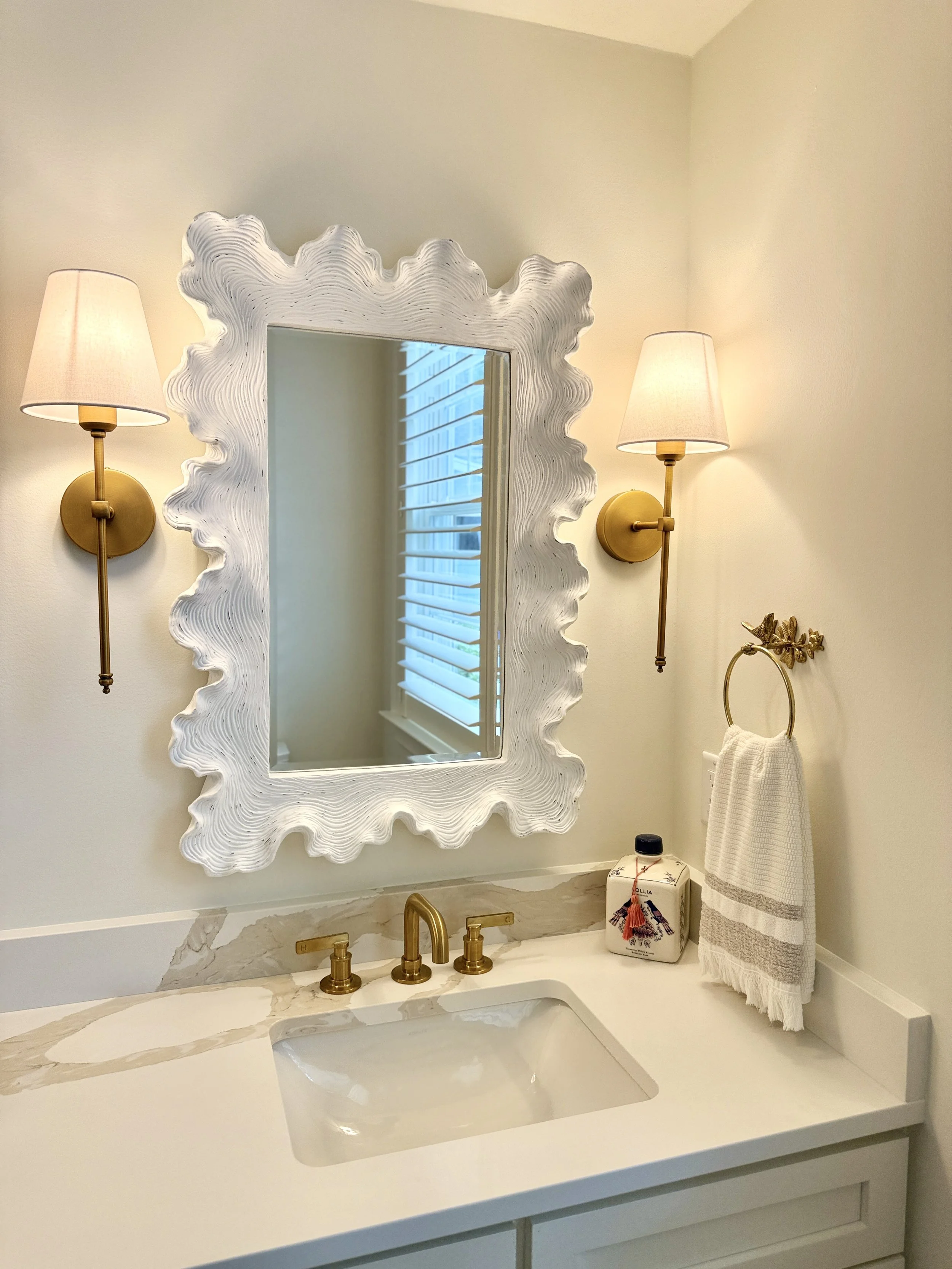 Bathroom with a white marble countertop, a rectangular sink, gold faucet and handles, a large white decorative mirror with a wavy frame, two wall-mounted gold lamps with beige shades, a gold towel ring with a white towel, a small decorative box with a flower and red tassel, and a window with white blinds.