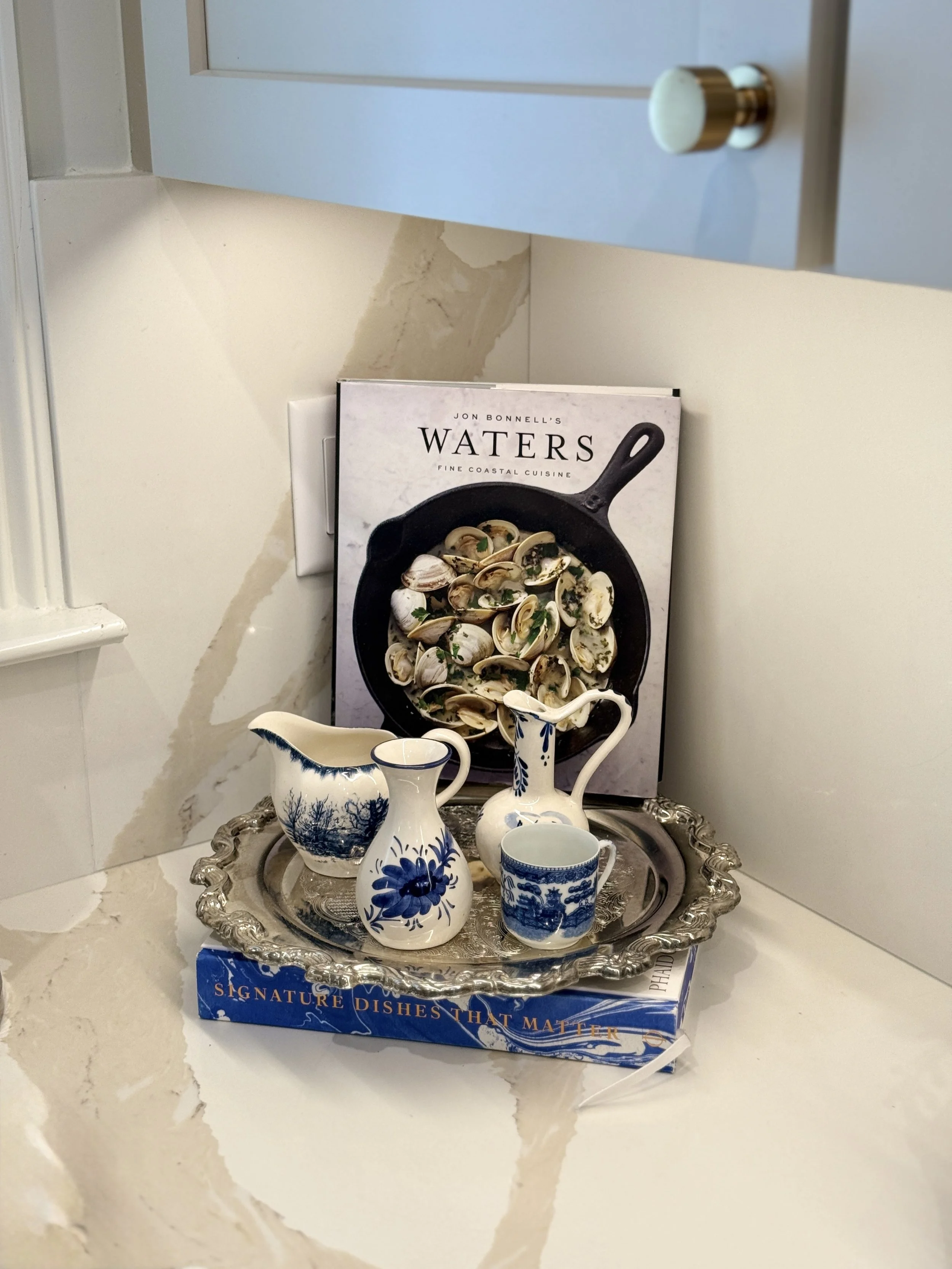 A decorative silver tray with white and blue porcelain pitchers, cups, and vases sitting on a marble countertop. There is a cookbook titled "Water" by Jon Bonnell behind the tray. The book features a black skillet with cooked clams on its cover.