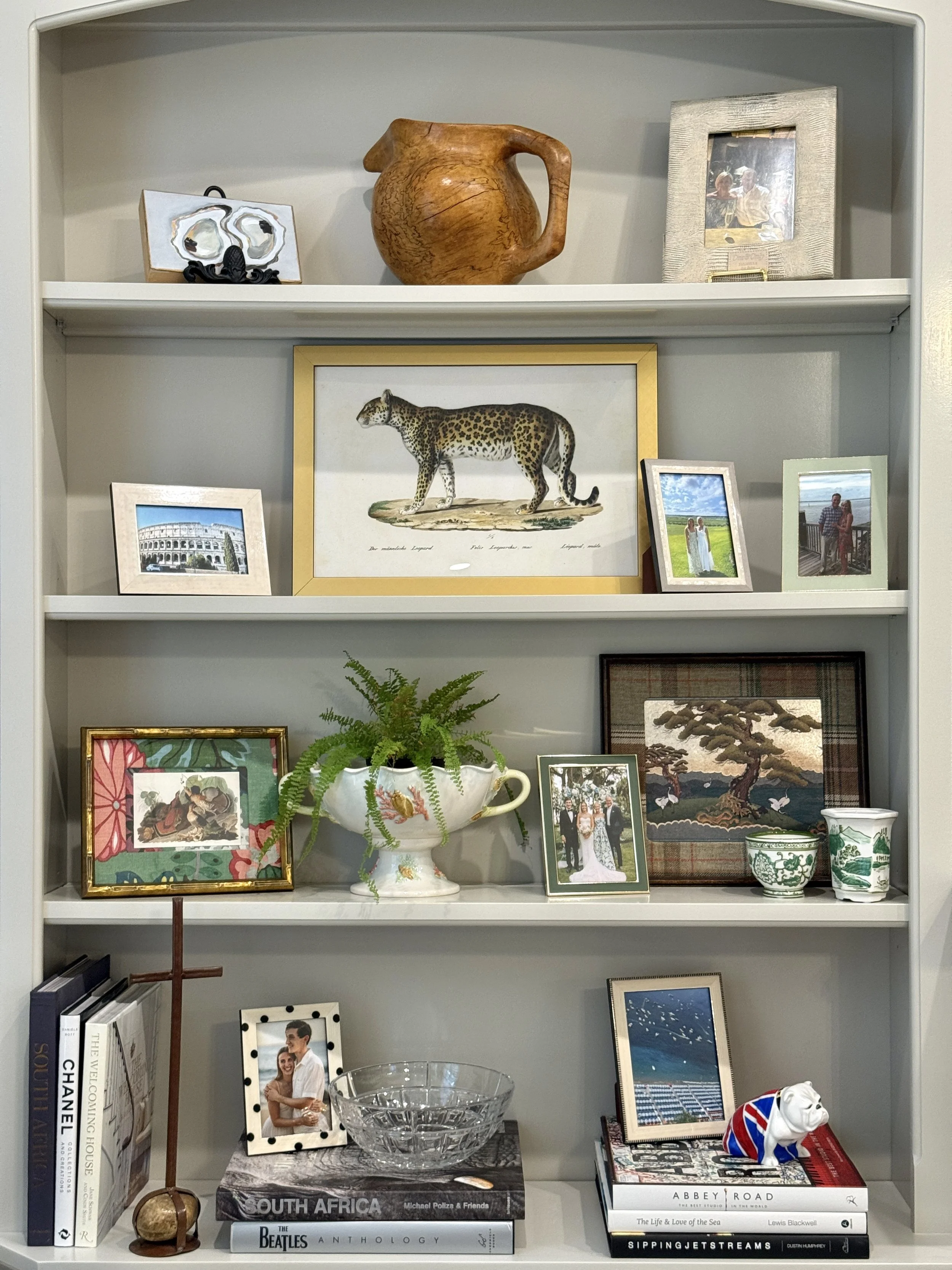 A white bookshelf displaying framed photographs, books, decorative items, and small plants, with a diverse collection of artwork and souvenirs.