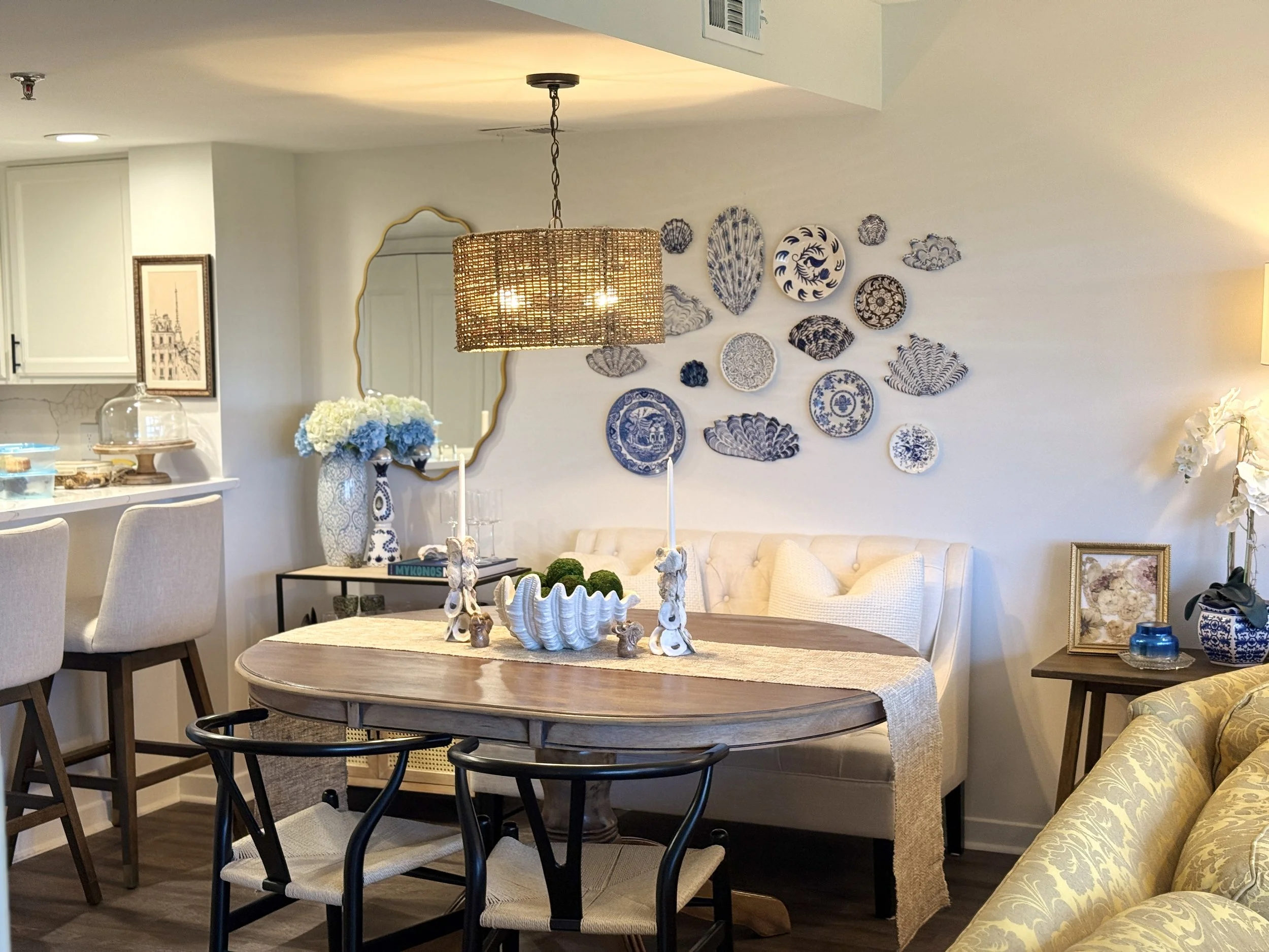 A cozy dining room and living space decorated with blue and white pottery, a rattan pendant light, a round wooden dining table with black and white chairs, and a wall art display of seashells and plates. There are floral arrangements, framed pictures