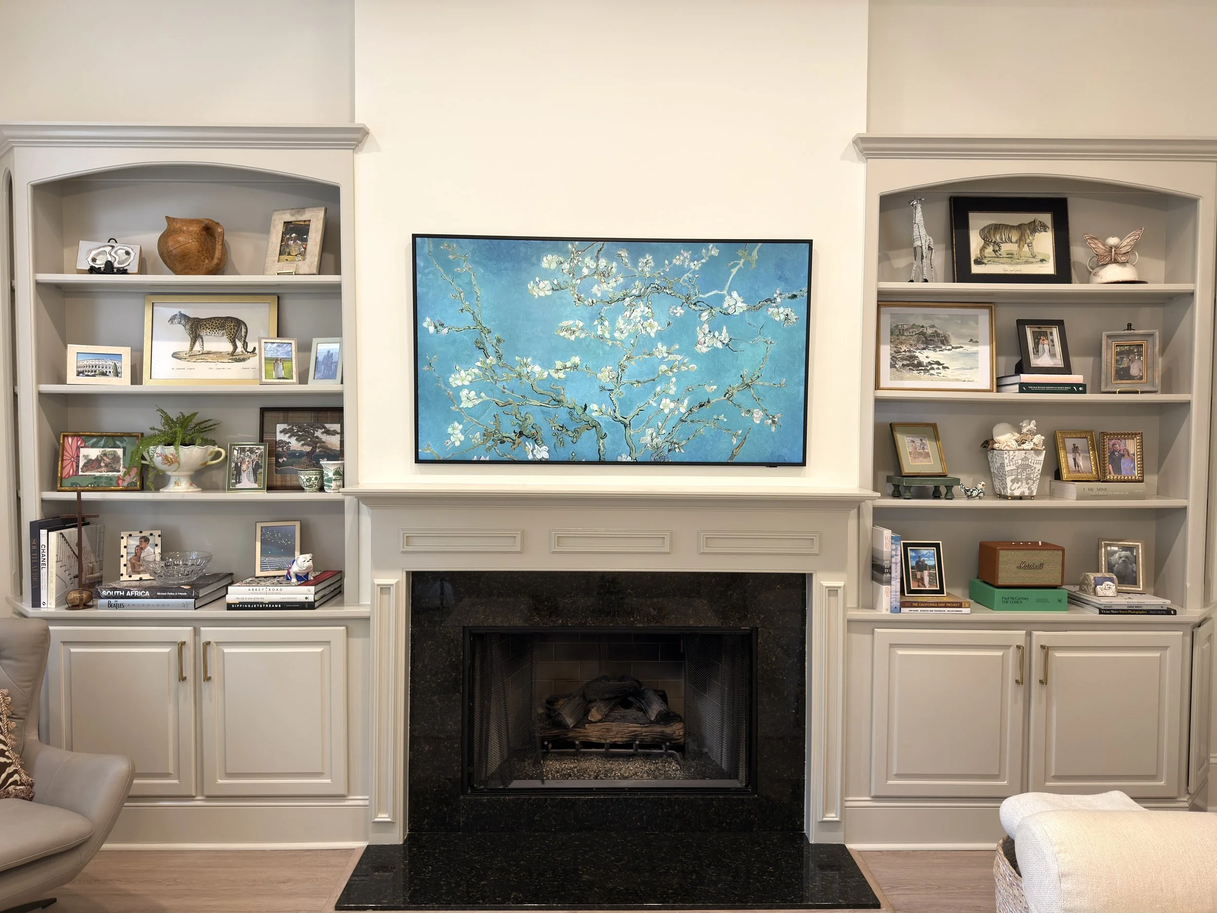 A living room fireplace with white wooden cabinets on either side, a flat-screen TV displaying a painting of white blossoms on a blue background above the fireplace, and various decorative items and framed photographs on the shelves.