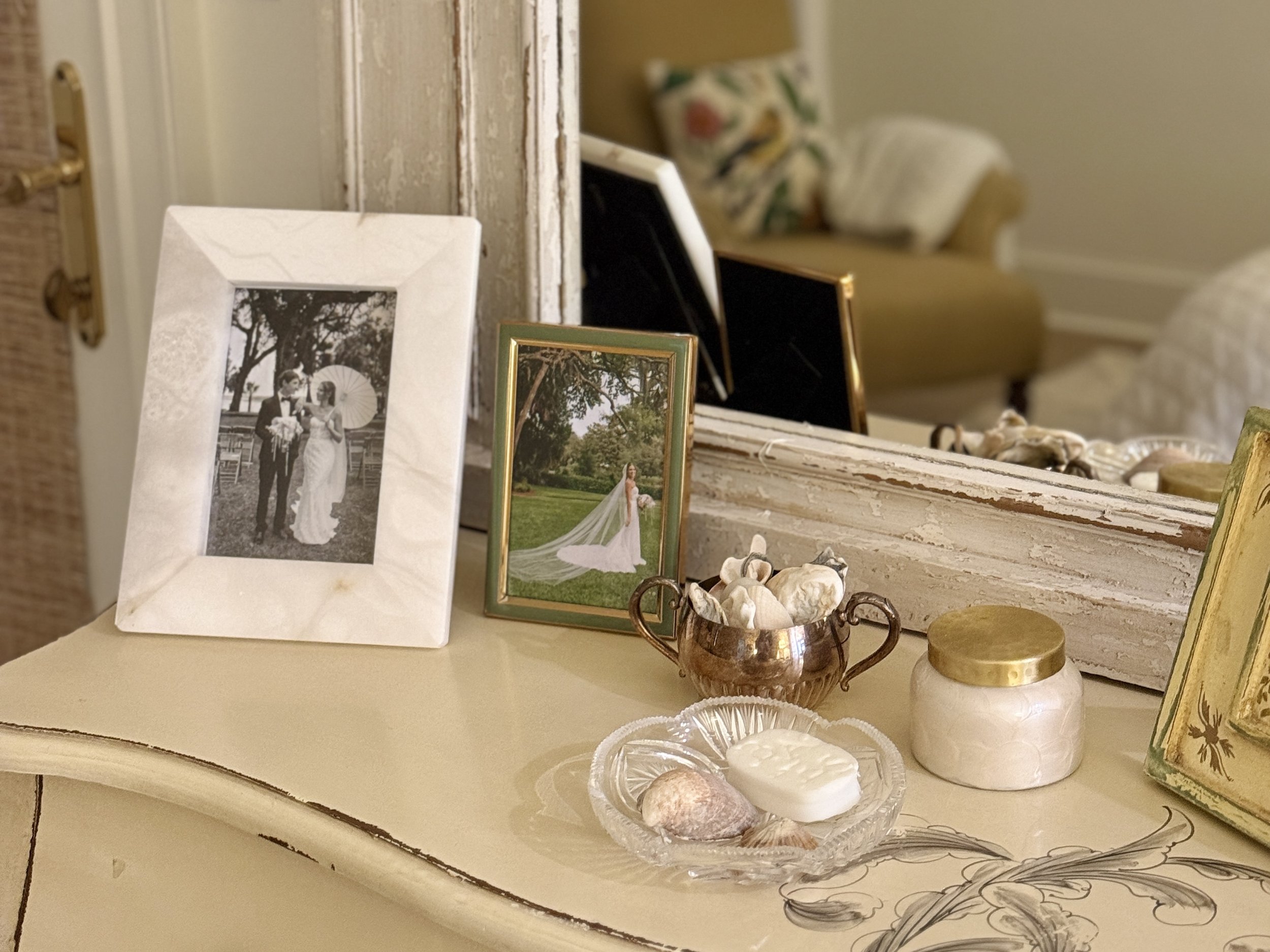 A beige dresser with framed photographs of a couple outdoors, a copper bowl with shells, a dish with seashells, and a decorative cream-colored jar.