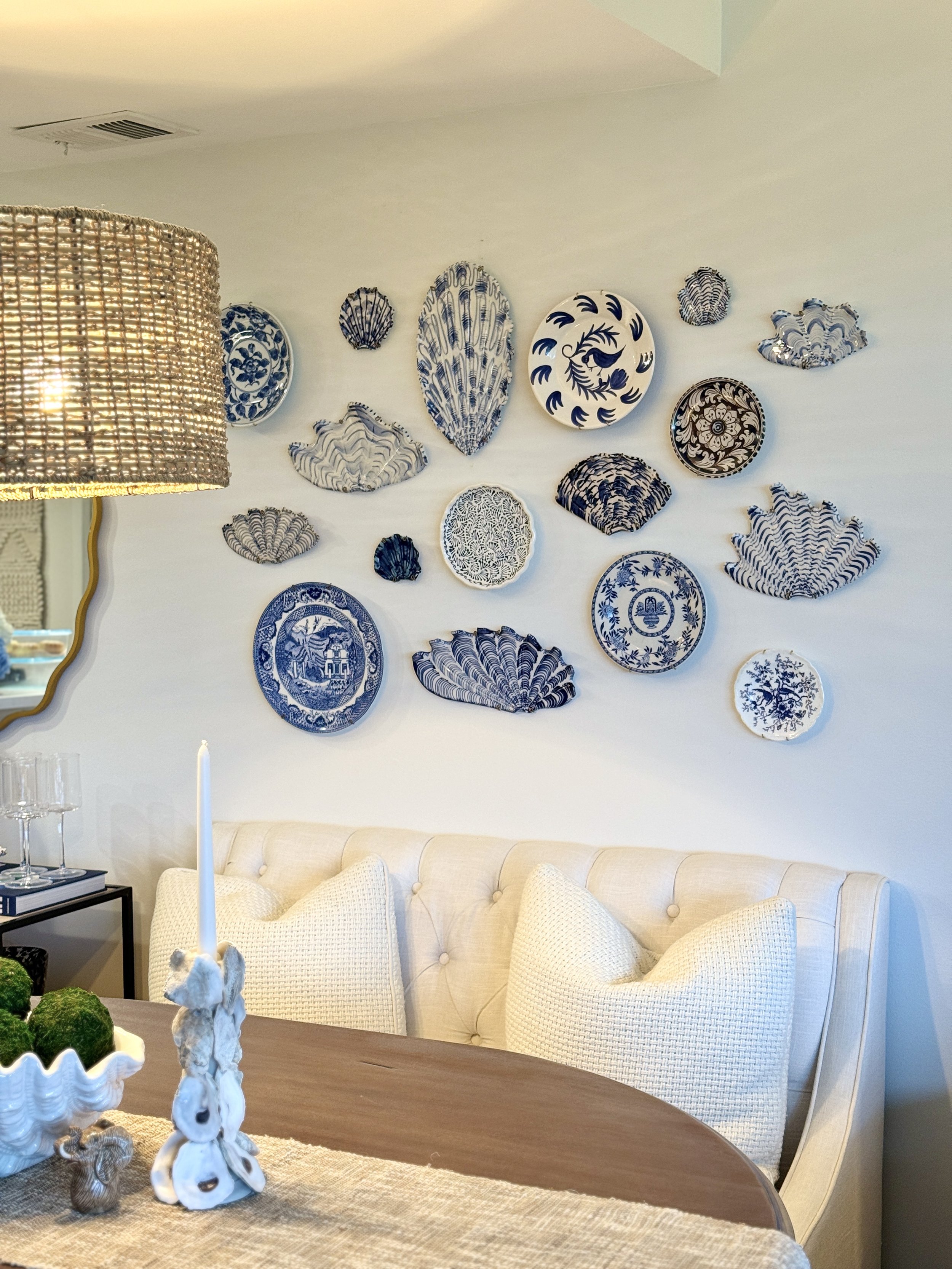 Decorative wall display of blue and white porcelain plates and shells above a cream-colored tufted sofa in a dining room with a wooden table and a patterned table runner.
