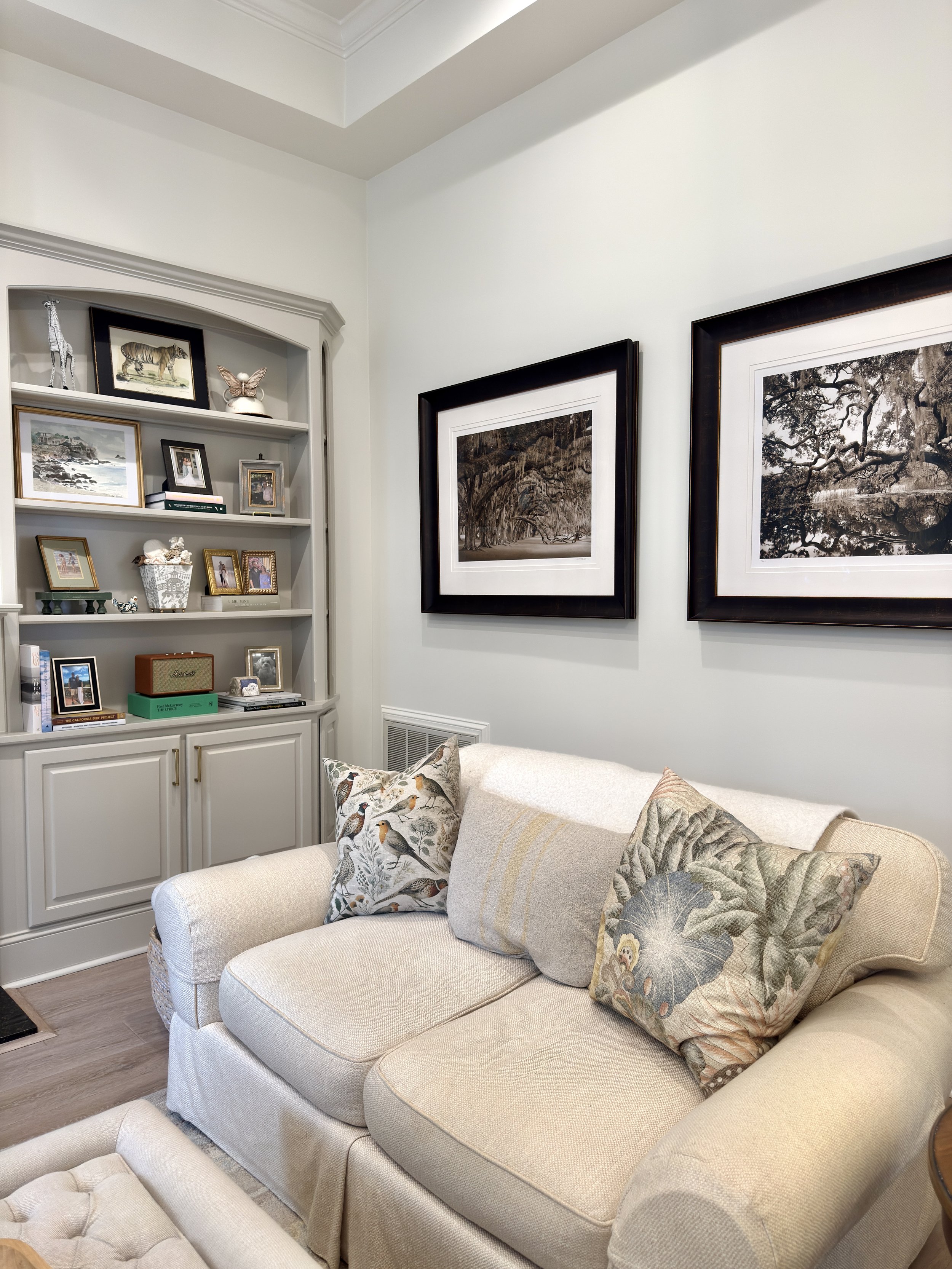 A living room featuring a beige upholstered sofa with decorative pillows, a cream-colored curvy armchair in the foreground, and a white built-in bookcase filled with framed family photos, books, and decorative items. Two black-framed landscape photog