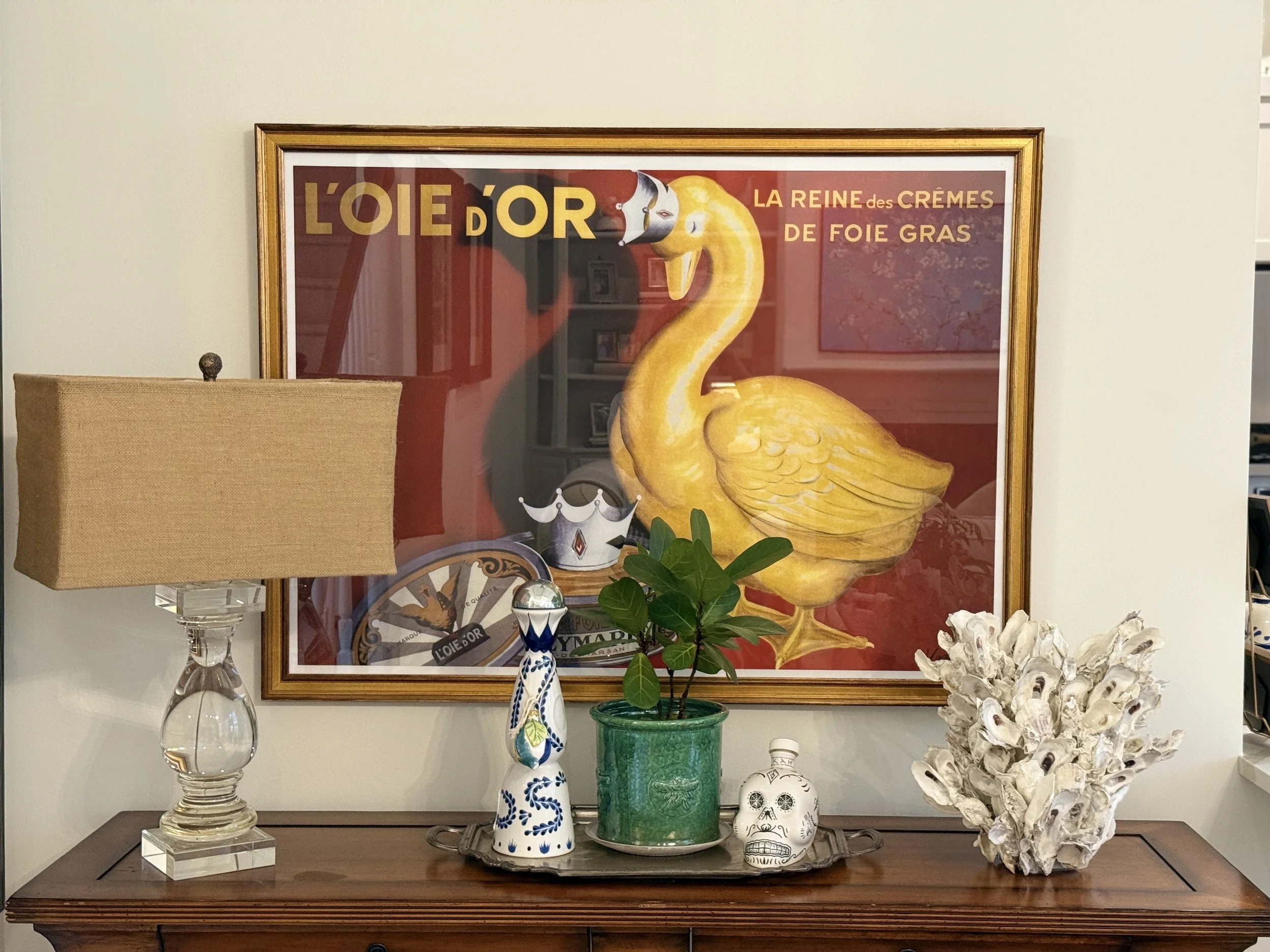 A wooden table with decorative items including glass lamps, decorated skulls, a potted plant, and a tray holding more objects. Behind the table, there is a colorful framed poster featuring a yellow swan with a crown and the text 'L'OIE D'OR La Reine 