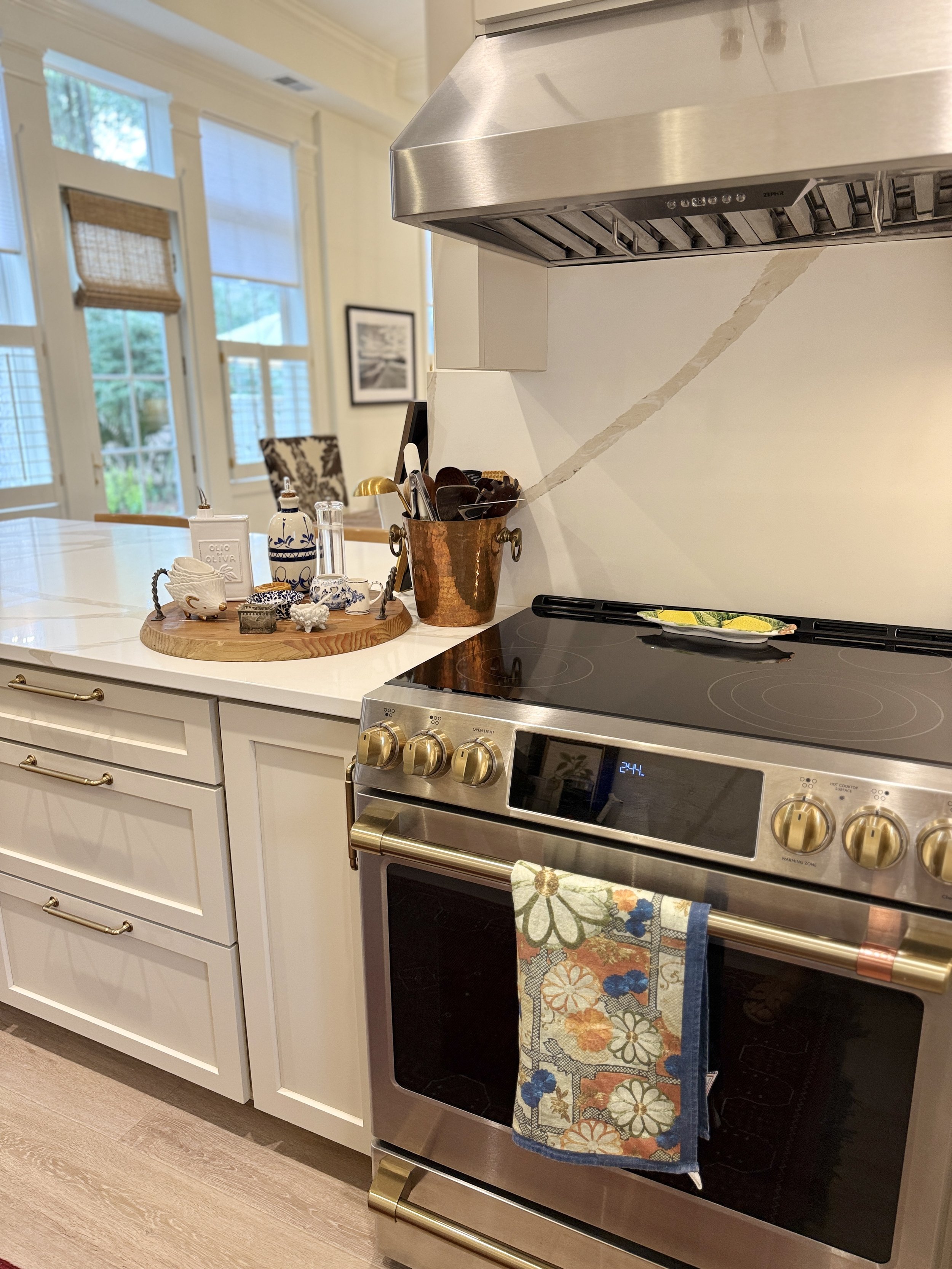 A modern kitchen with white cabinets, a black cooktop, a stainless steel oven, and a decorative towel hanging from the oven handle. There are various kitchen items on a wooden tray and a copper container on the countertop. In the background, large wi