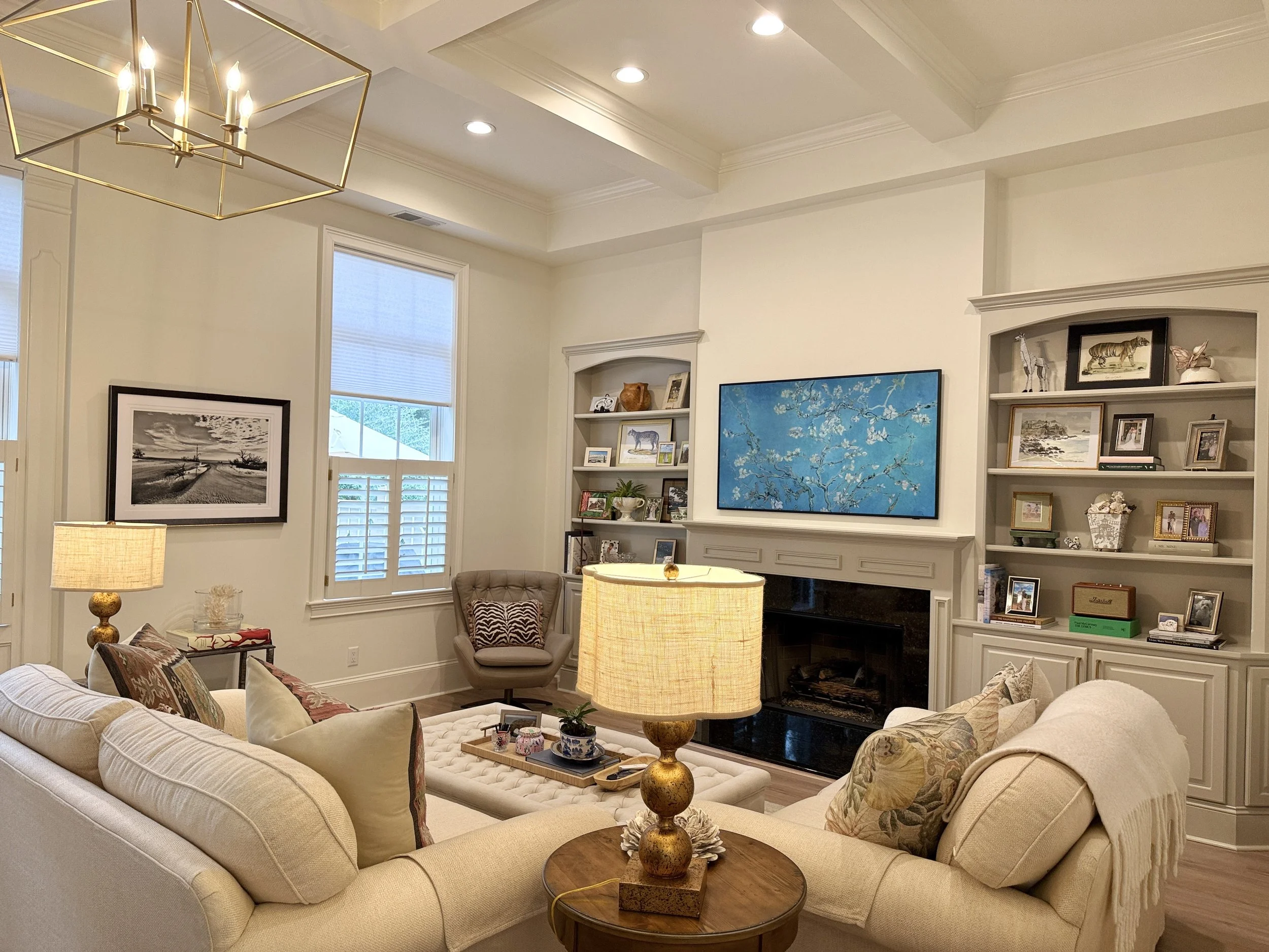 Living room with beige sofas and armchair, wooden coffee table, lamps, built-in white bookshelves with framed photos and artwork, a blue floral painting above the fireplace, and windows with white blinds.
