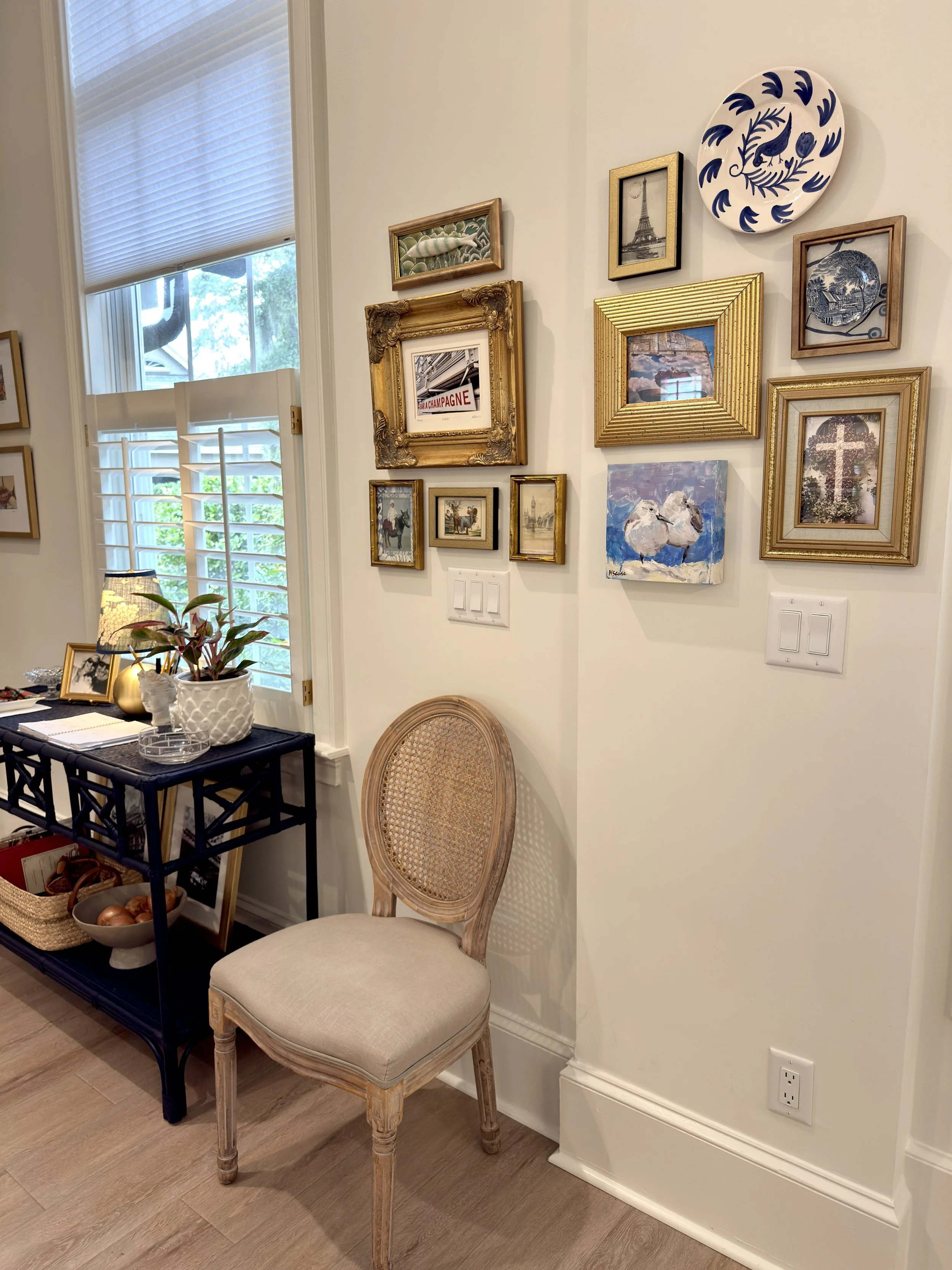 Corner of a room with wall decorated with framed pictures and plates, a beige chair, and a black side table with plants and decorative items, next to a window with blinds.