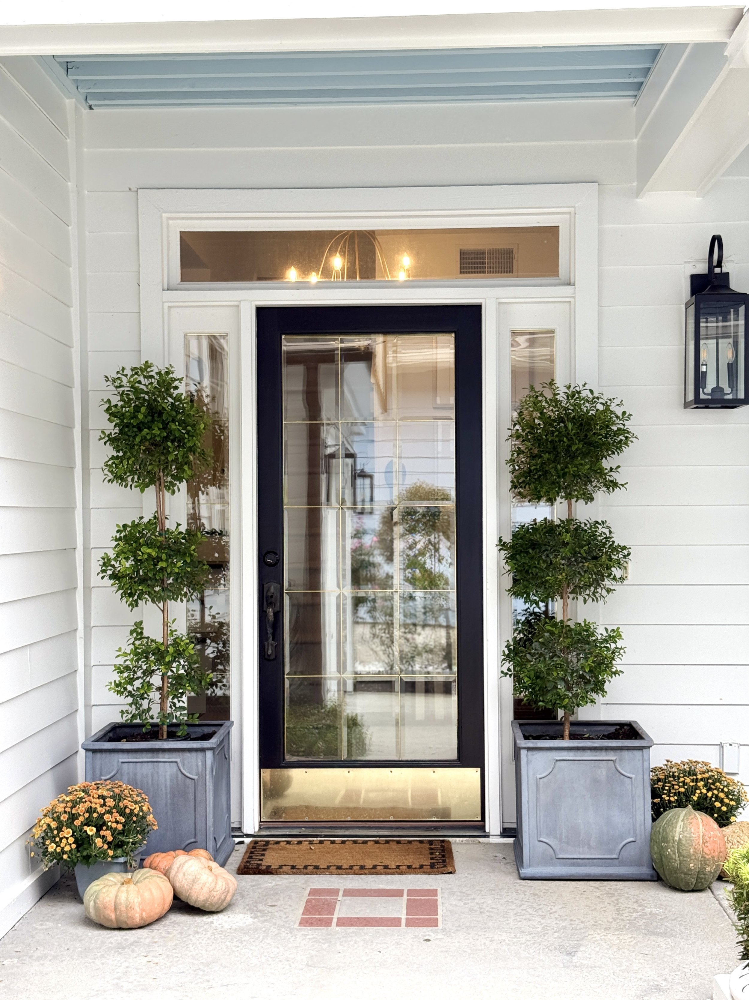 Front porch of a house decorated for autumn with pumpkins, mums, and potted plants, featuring a black front door with glass panes, flanked by two large planters with trimmed bushes and a vintage-style lantern on the wall.