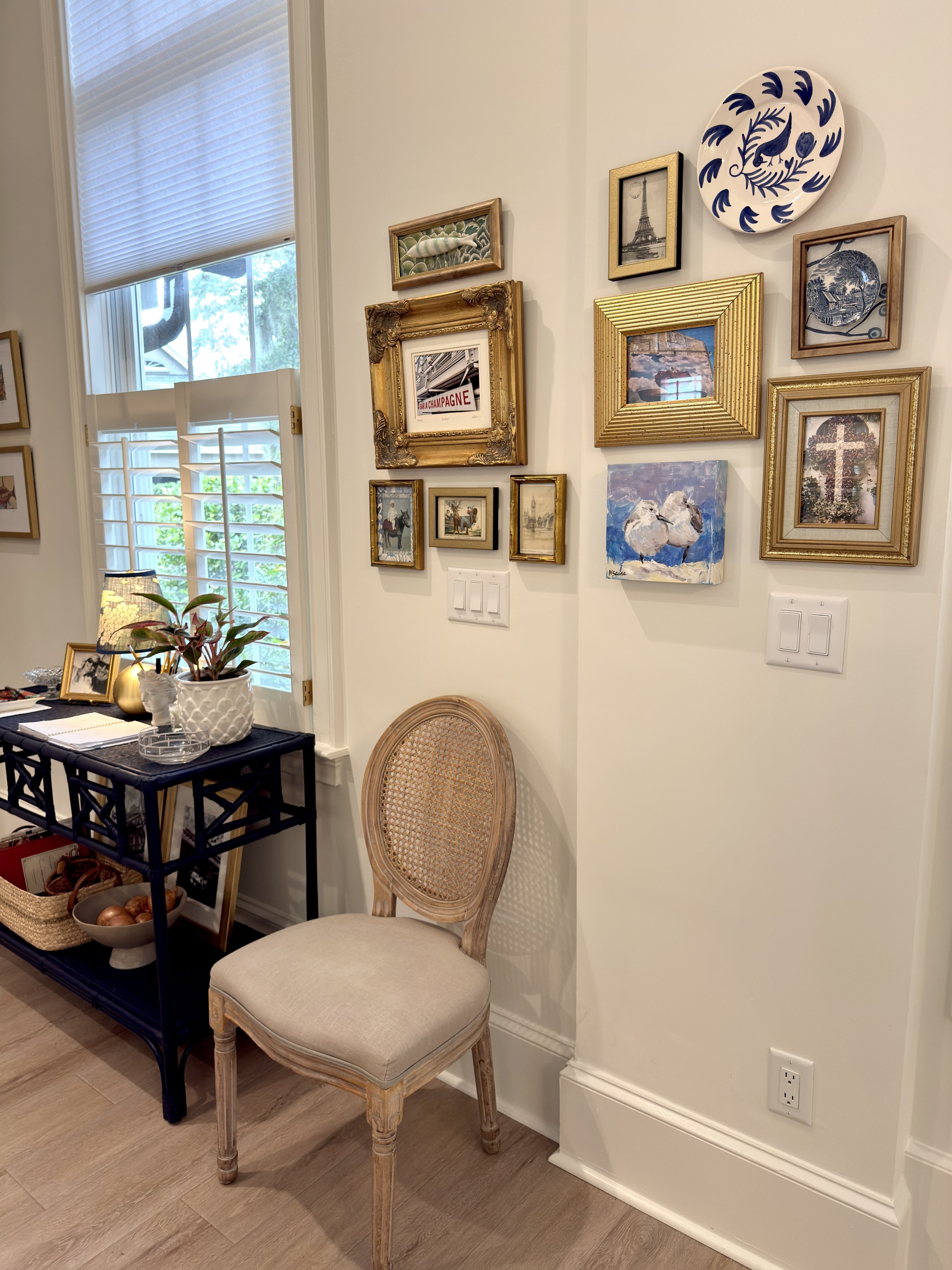 A cozy corner of a room with a vintage wooden chair with beige upholstery, a small black side table decorated with a potted plant, framed photos, and a bowl of onions. The wall features a gallery of framed art including paintings and photographs, wit