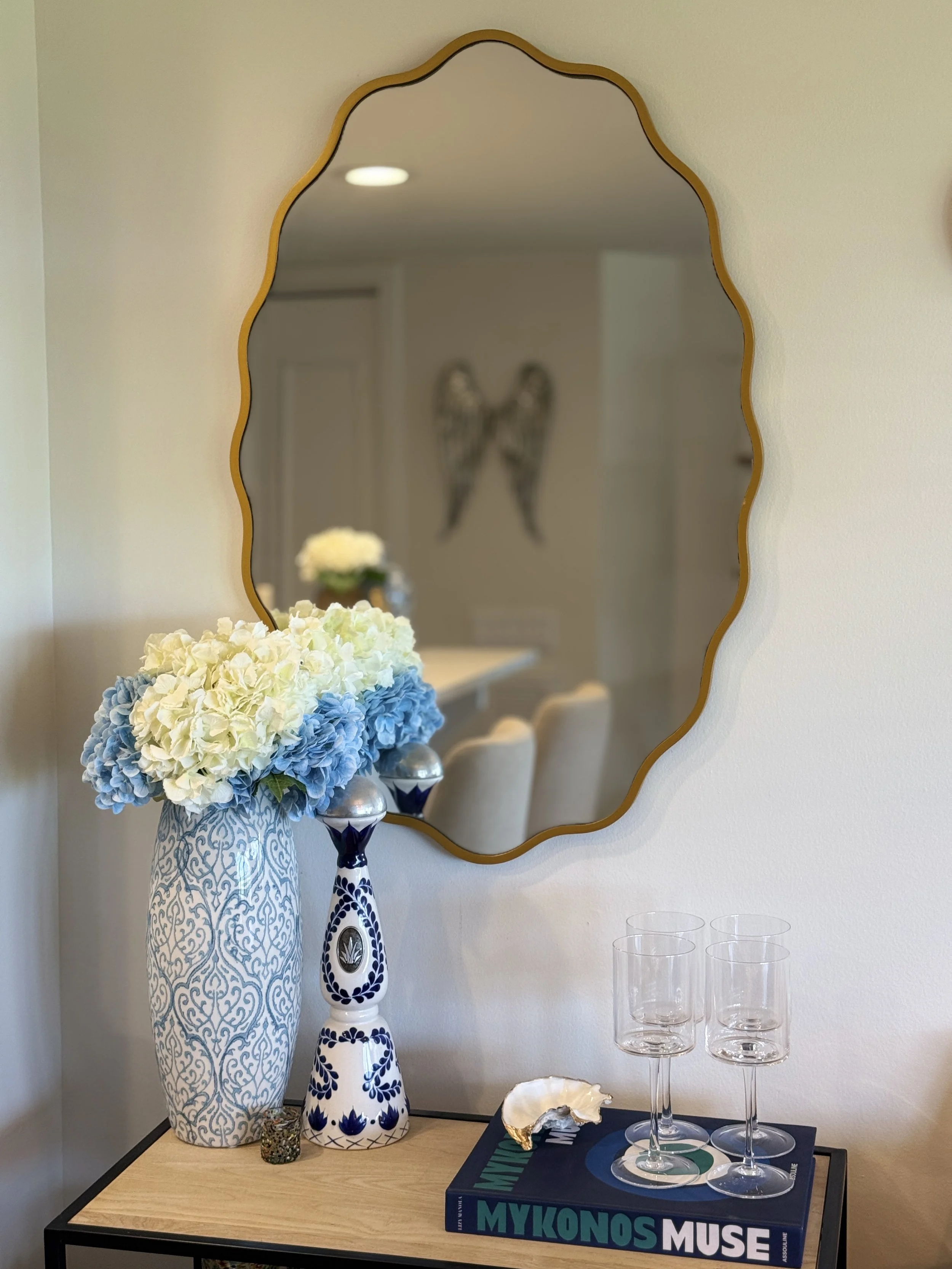 A decorative table arrangement includes a large vase with a blue and white pattern filled with white and light blue hydrangeas, a shorter vase with a similar pattern, a tall black and white vase, a large gold-framed mirror with a scalloped edge, three empty wine glasses, a decorative oyster shell, and a large hardcover book titled "Mykonos Muse," on a wooden table with a black metal frame in a room with beige walls.