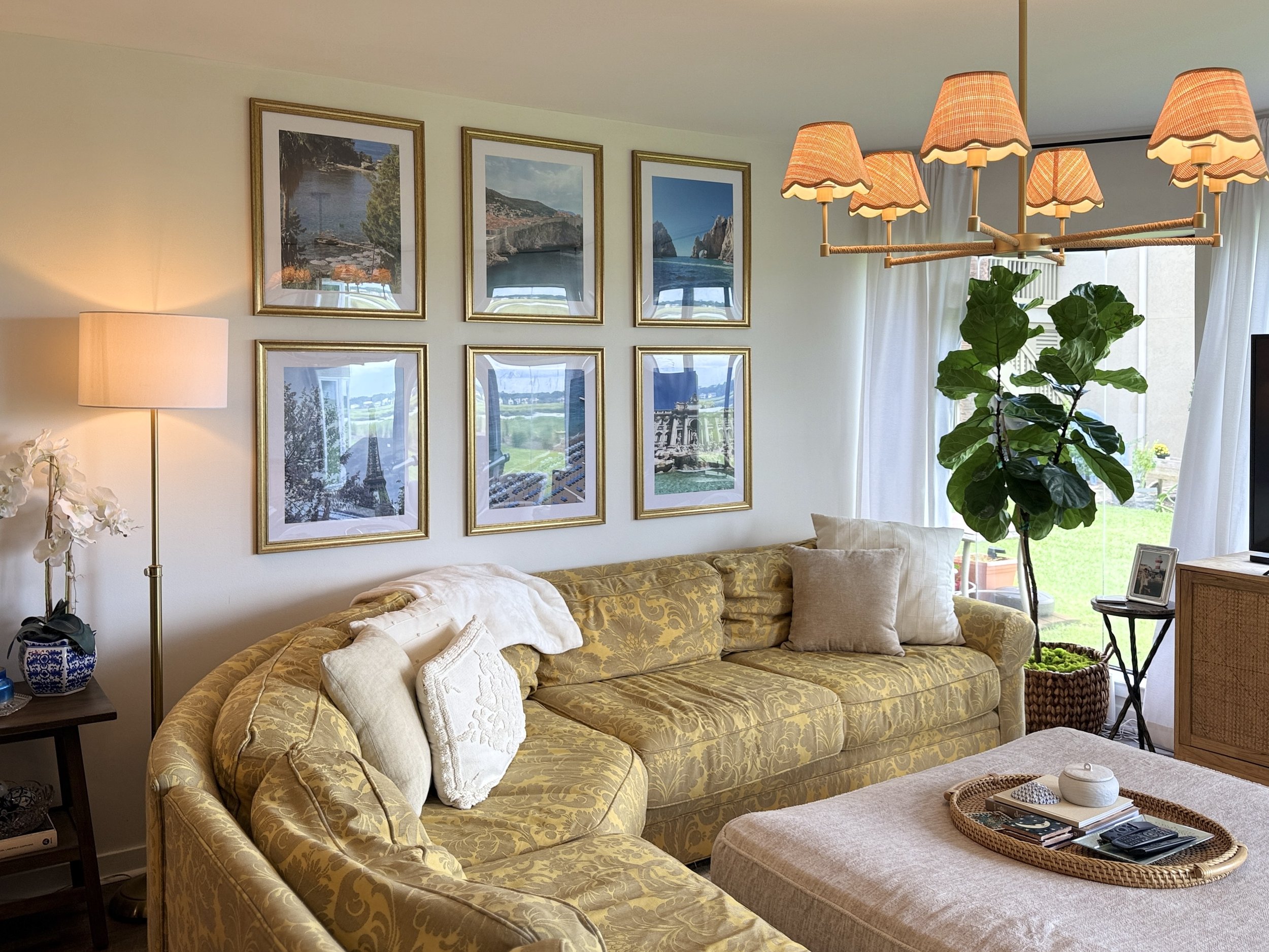Living room with yellow patterned sofa, white pillows, glass coffee table with tray, books, and remote, wall decorated with three large framed photographs of coastal landscapes, tall floor lamp with beige shade on left, large leafy green plant near w