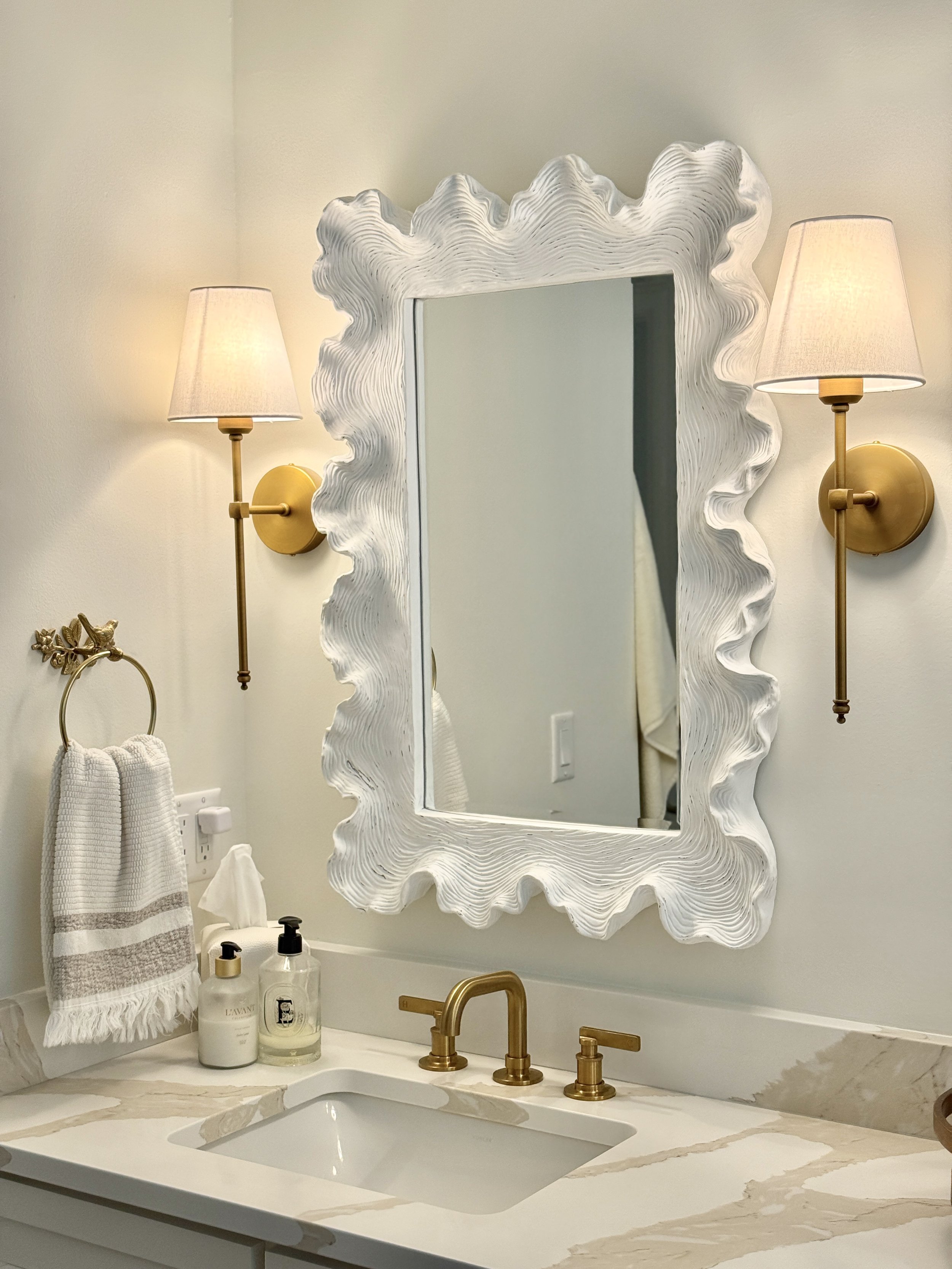 A bathroom sink with a white marble countertop, gold fixtures, and a decorative mirror with a wavy white frame. Two wall-mounted lamps with beige shades flank the mirror. On the counter are soap dispensers and a small gold decorative item. A towel ha
