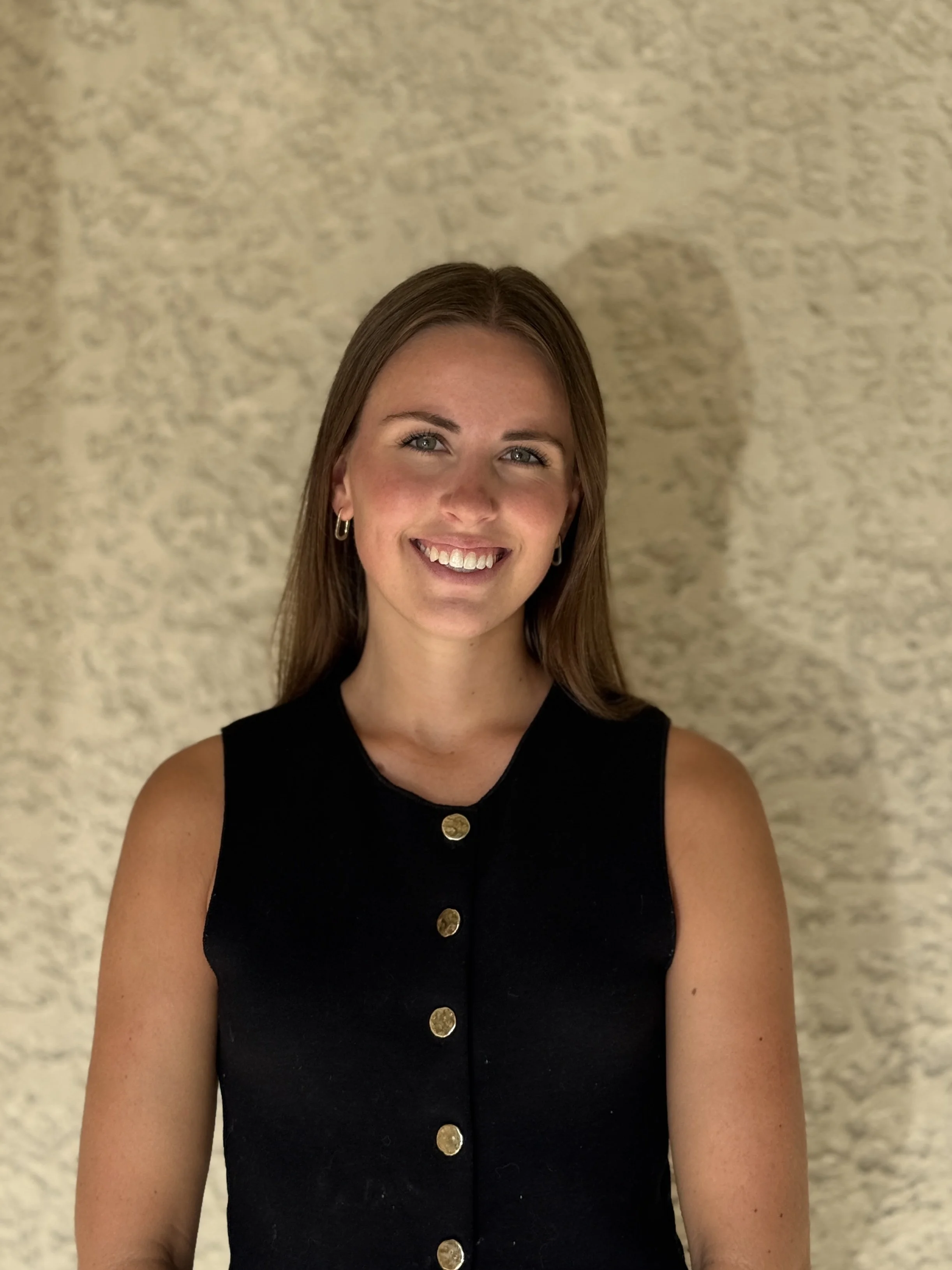 A woman with long brown hair, wearing a sleeveless black dress with gold buttons, smiling, standing against a textured beige wall.