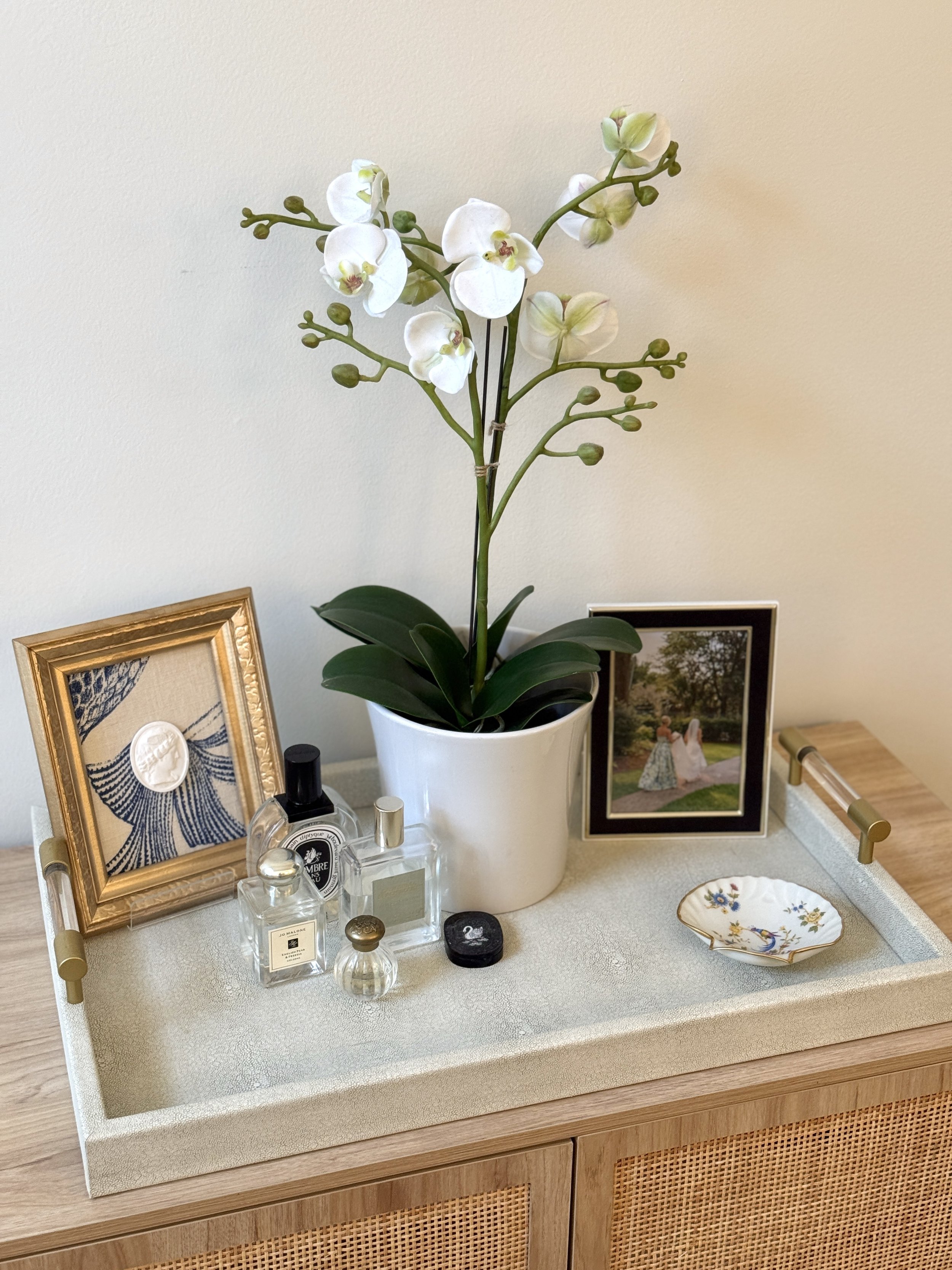 A potted white orchid on a white display table, decorated with framed photographs, perfume bottles, and a decorative dish.