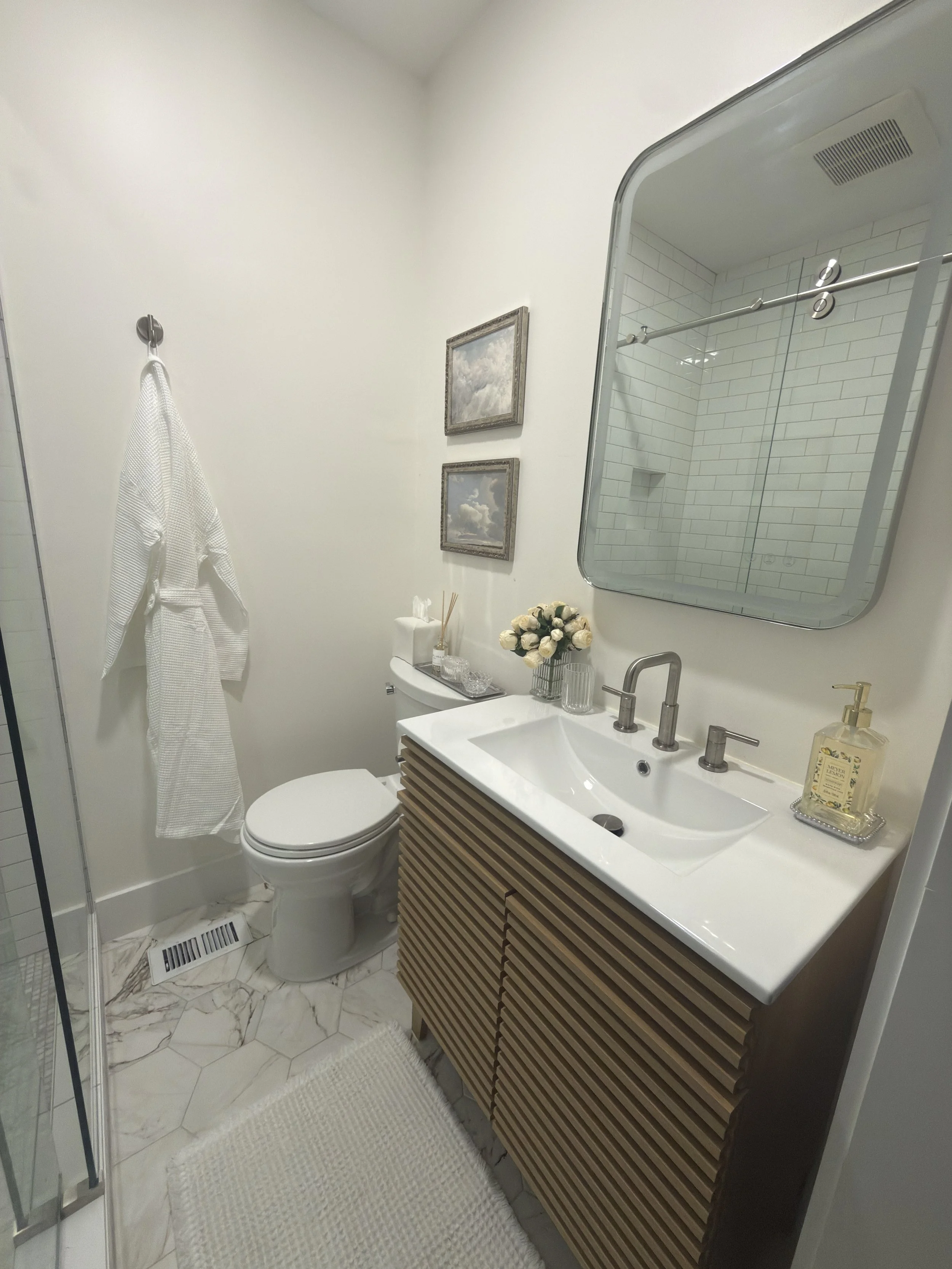 Bathroom with white walls, a white toilet, a wooden vanity with a white countertop and modern faucet, and a large mirror. Decor includes framed artwork, a vase with white roses, and hand soap. There's a shower with glass doors, white subway tile, and