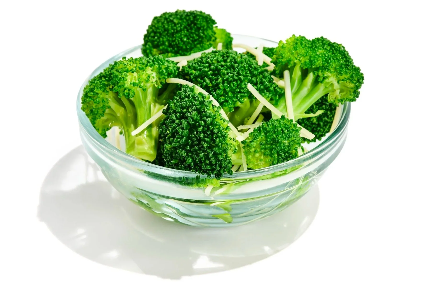 Fresh broccoli florets in a clear glass bowl with shredded cheese on top.
