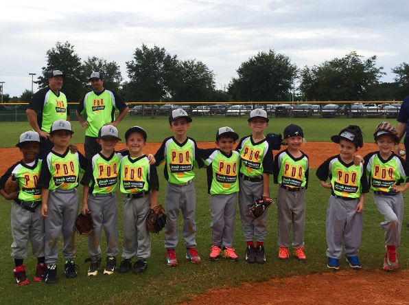A group of young children wearing matching baseball uniforms and caps standing in a line on a baseball field. Two adults in similar uniforms are standing behind them. Trees and a fence with cars in the background. It appears to be a team photo.