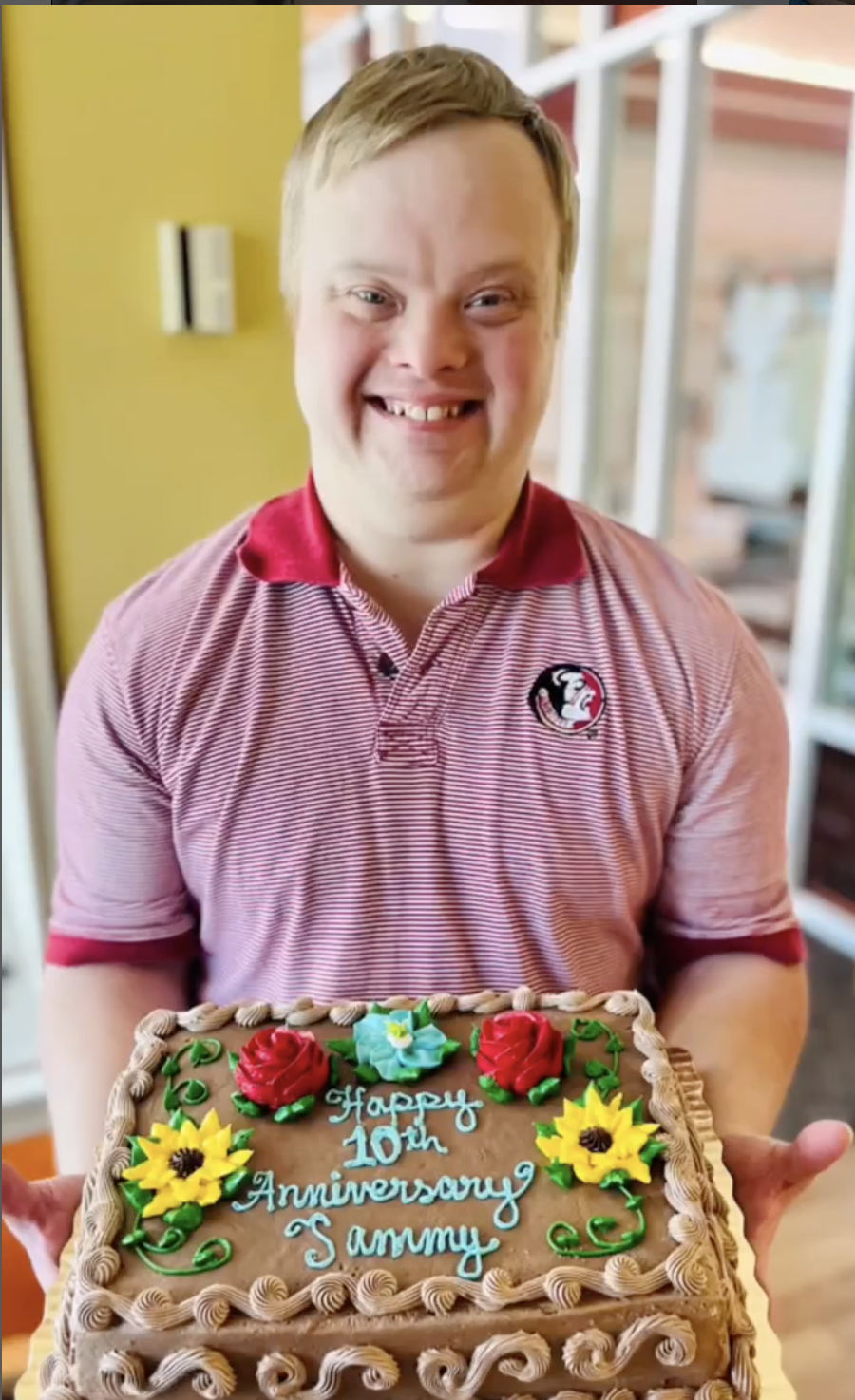 Person holding a decorated cake with the text 'Happy 10th Anniversary Sammy' and floral designs, wearing a striped shirt.