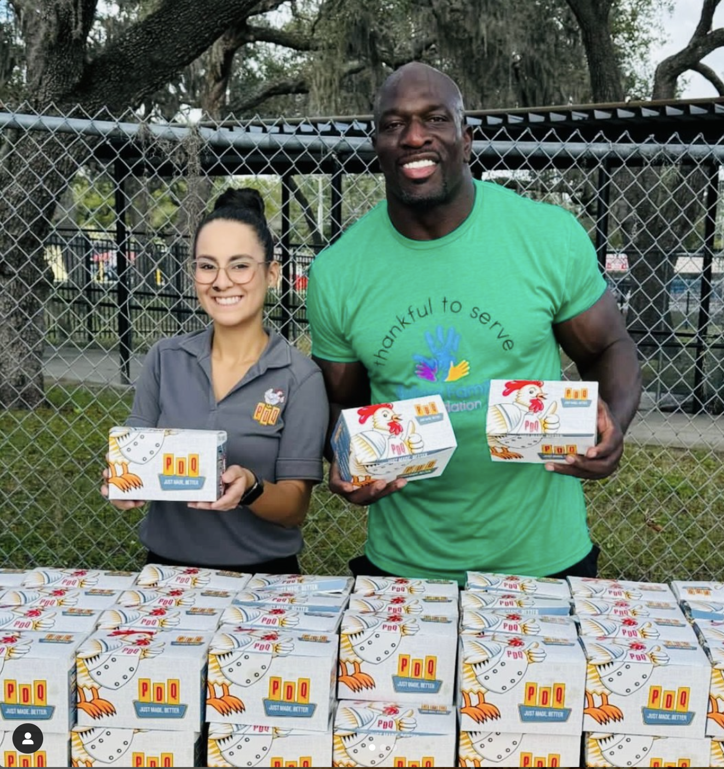 Two people smiling and holding PDQ food boxes in front of a table with more PDQ boxes, outdoors near a metal fence.
