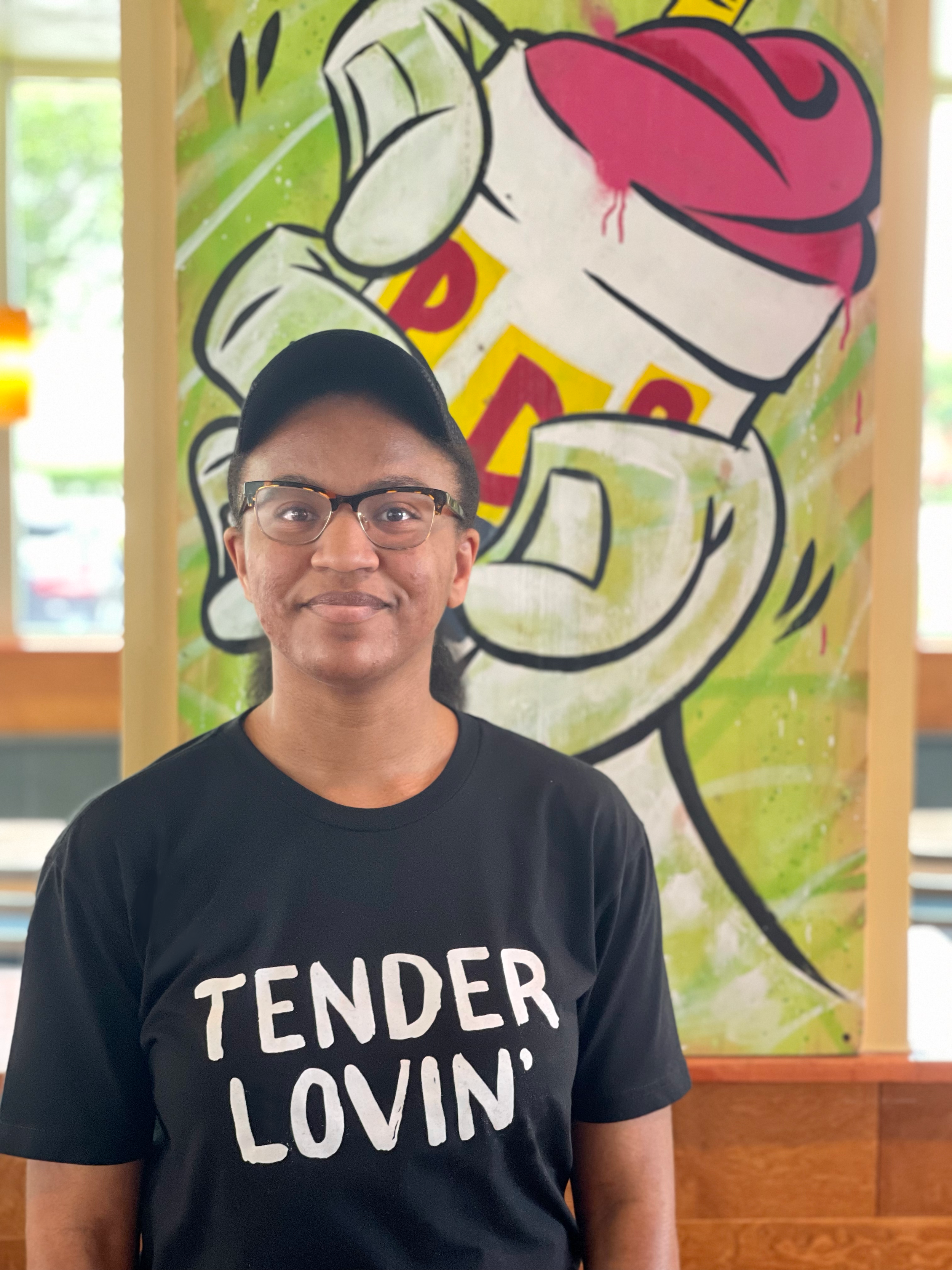 Person wearing glasses and a black "Tender Lovin'" t-shirt stands in front of a colorful mural of a cup with the word "RADS" on it.
