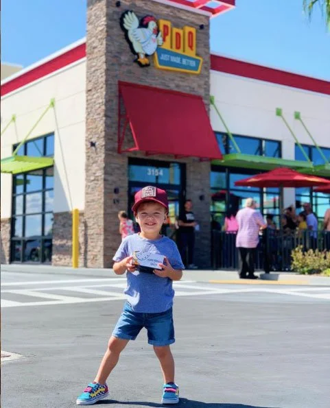 A child in casual attire stands smiling in front of a PDQ restaurant, holding a food container. The building features a large logo with a chicken above the entrance and vibrant red and green awnings. Several people are dining outdoors under umbrellas.