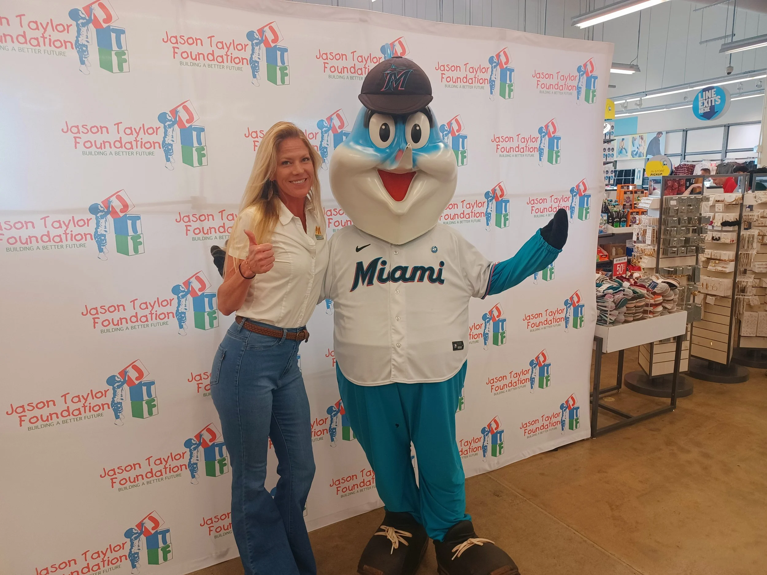 A person posing with a mascot in a Miami Marlins jersey and hat at a promotional event for the Jason Taylor Foundation. The backdrop has the foundation's logo.