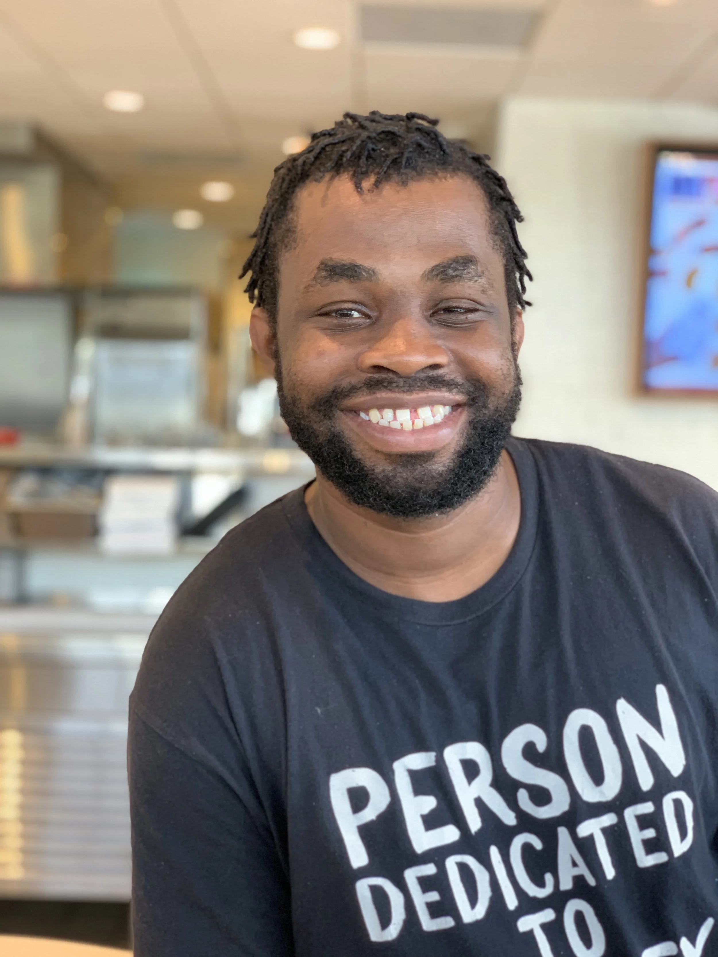 Smiling person wearing a black t-shirt with "PERSON DEDICATED TO X" text in a bright room.