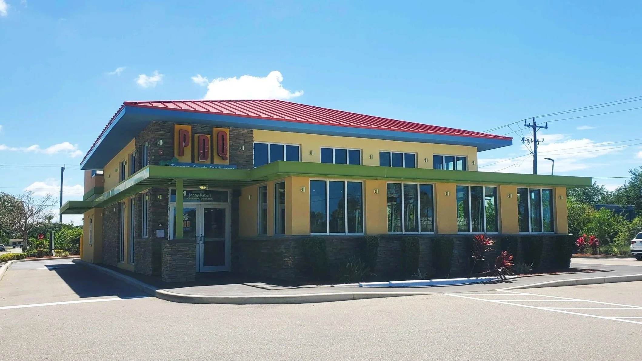 A modern two-story building with yellow walls, large glass windows, and a red roof. There are green accents on the sides and a stone facade near the entrance. The building is in a parking lot with a few cars and landscaping with bushes and small plants. Power lines are visible against a blue sky with some clouds.