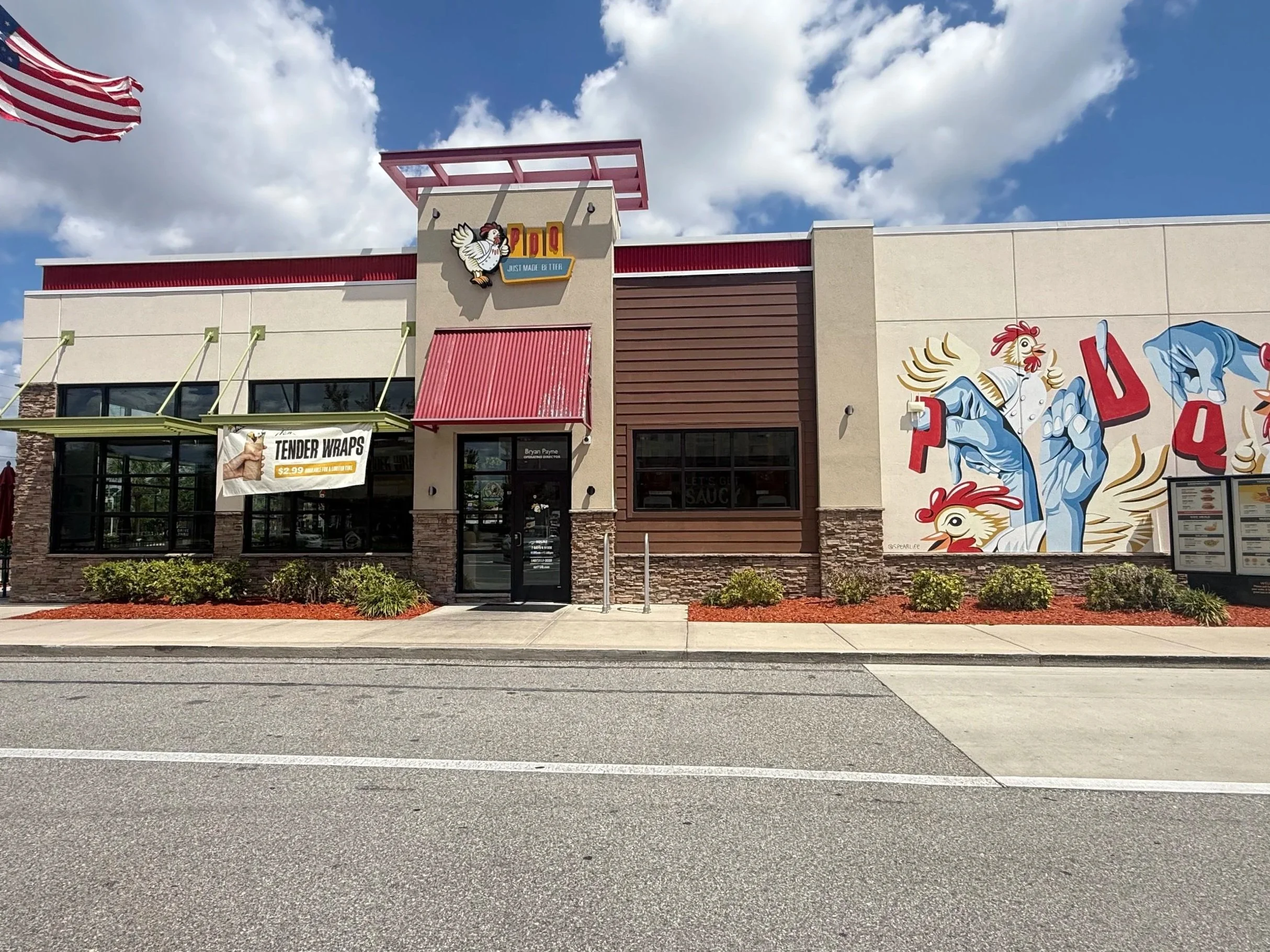 Front view of a fast food restaurant with a colorful mural on the side wall, a red awning over the entrance, a banner advertising tender wraps, and an American flag flying in the wind, under a partly cloudy sky.