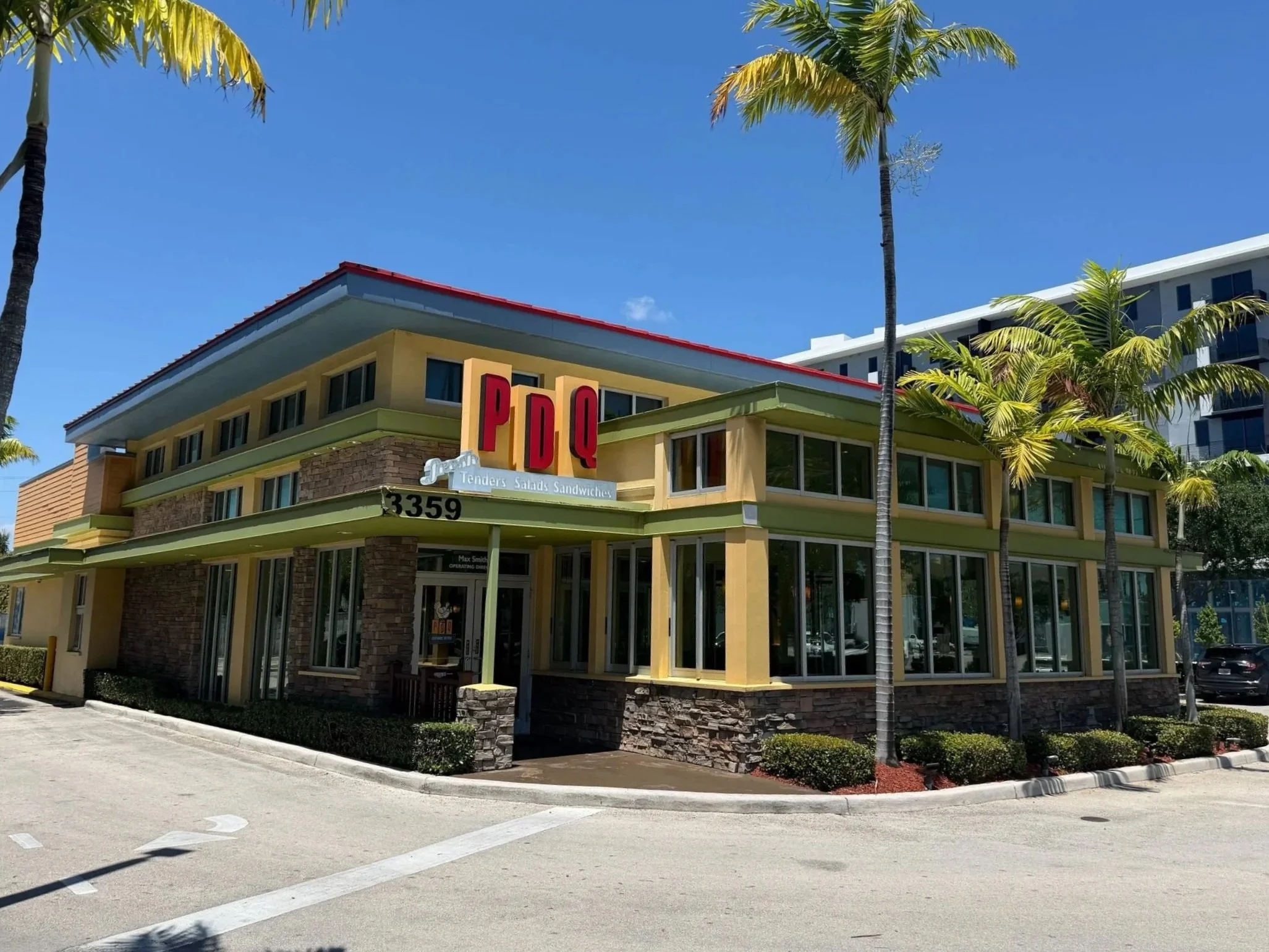 A colorful fast-food restaurant building with palm trees in front, a blue sky above, and street parking.