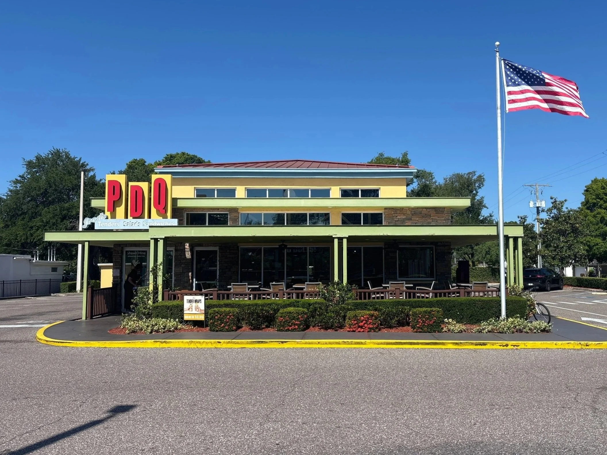 A fast food restaurant building with a yellow and green exterior, a red roof, and large red sign reading 'PDQ'. An American flag is flying on a tall flagpole in front of the building. There are trees, utility poles, and clear blue sky in the background.