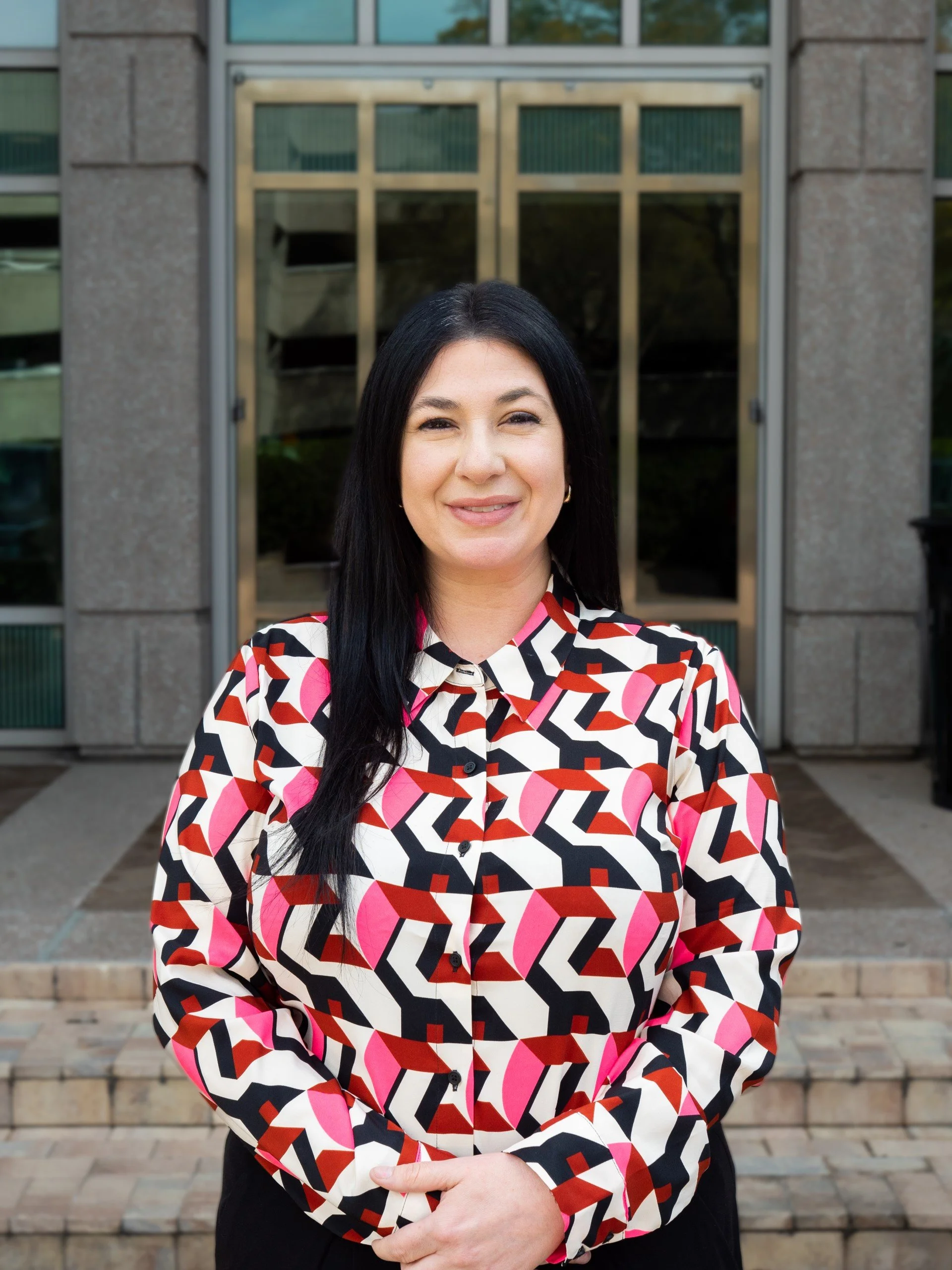 Woman with long dark hair wearing a colorful geometric shirt, smiling, standing in front of a building entrance with glass doors.