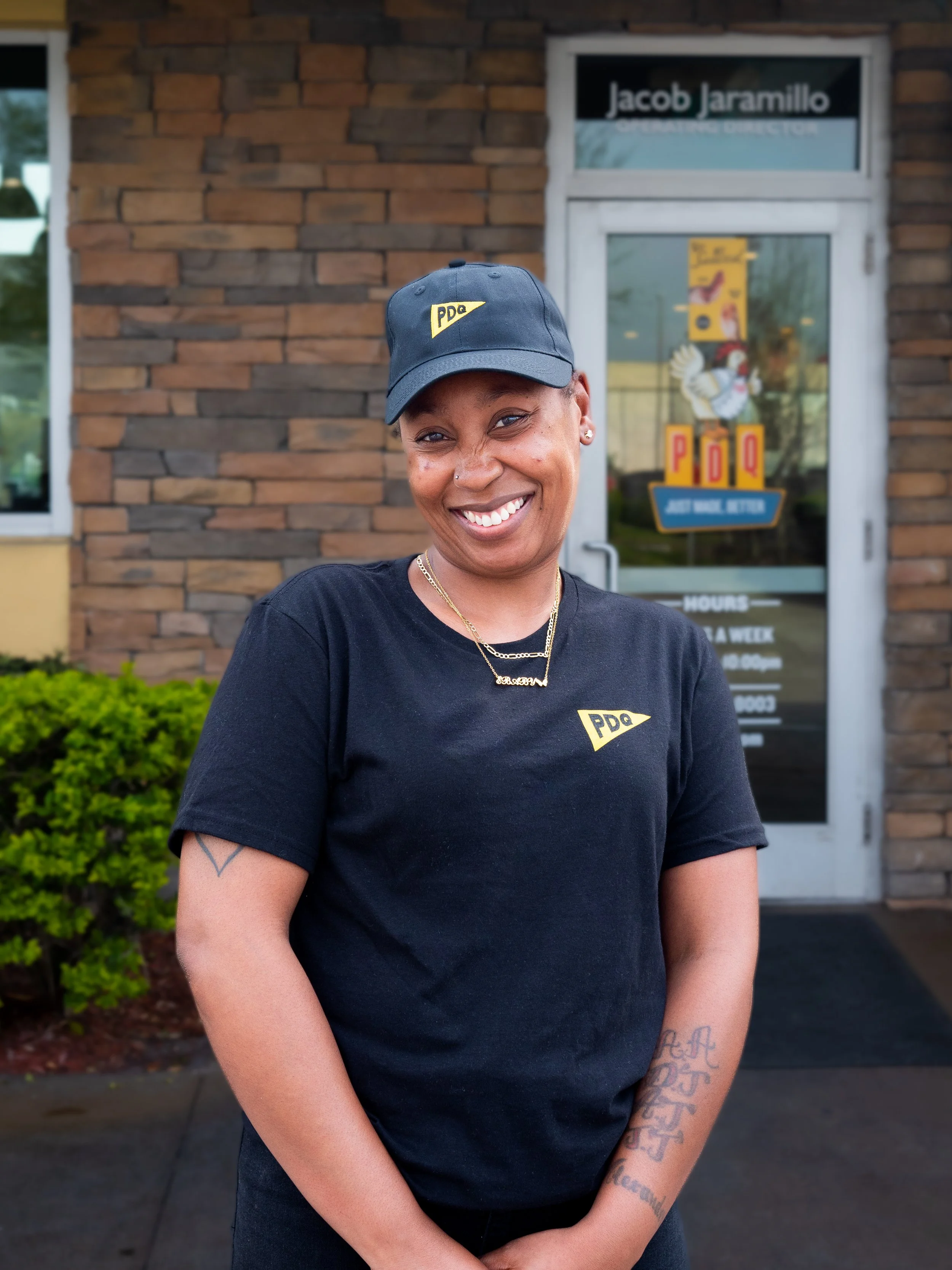 Smiling team member in black PDQ uniform standing outside a brick building with a glass door.