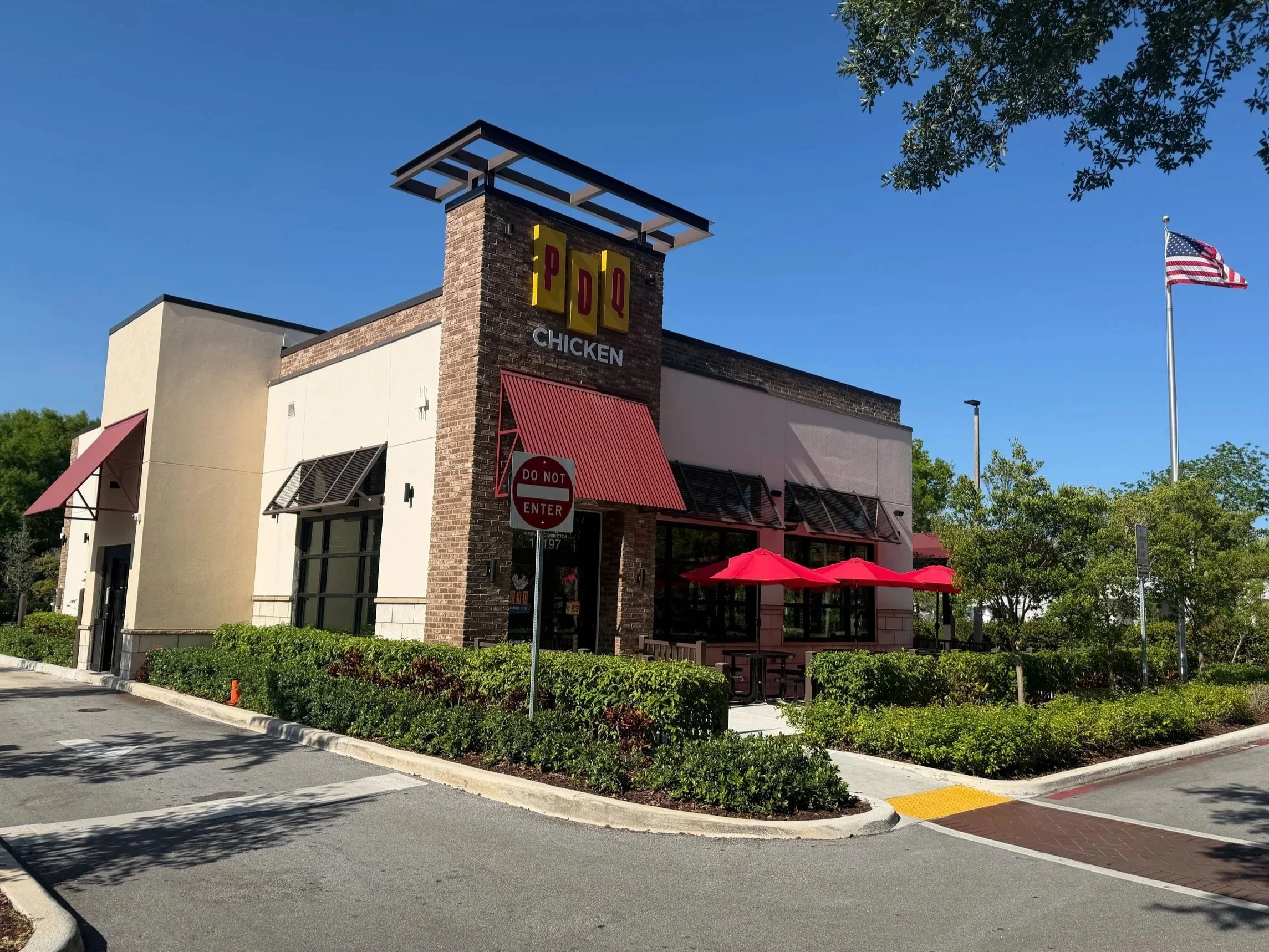 A fast food restaurant called PDQ with a sign that says 'Chicken'. The building has a brick and beige exterior with red awnings and outdoor seating with red umbrellas. There are green bushes, a 'Do Not Enter' sign, and a parking lot in front. An American flag is flying on a flagpole, and trees are visible in the background. The sky is clear and blue.