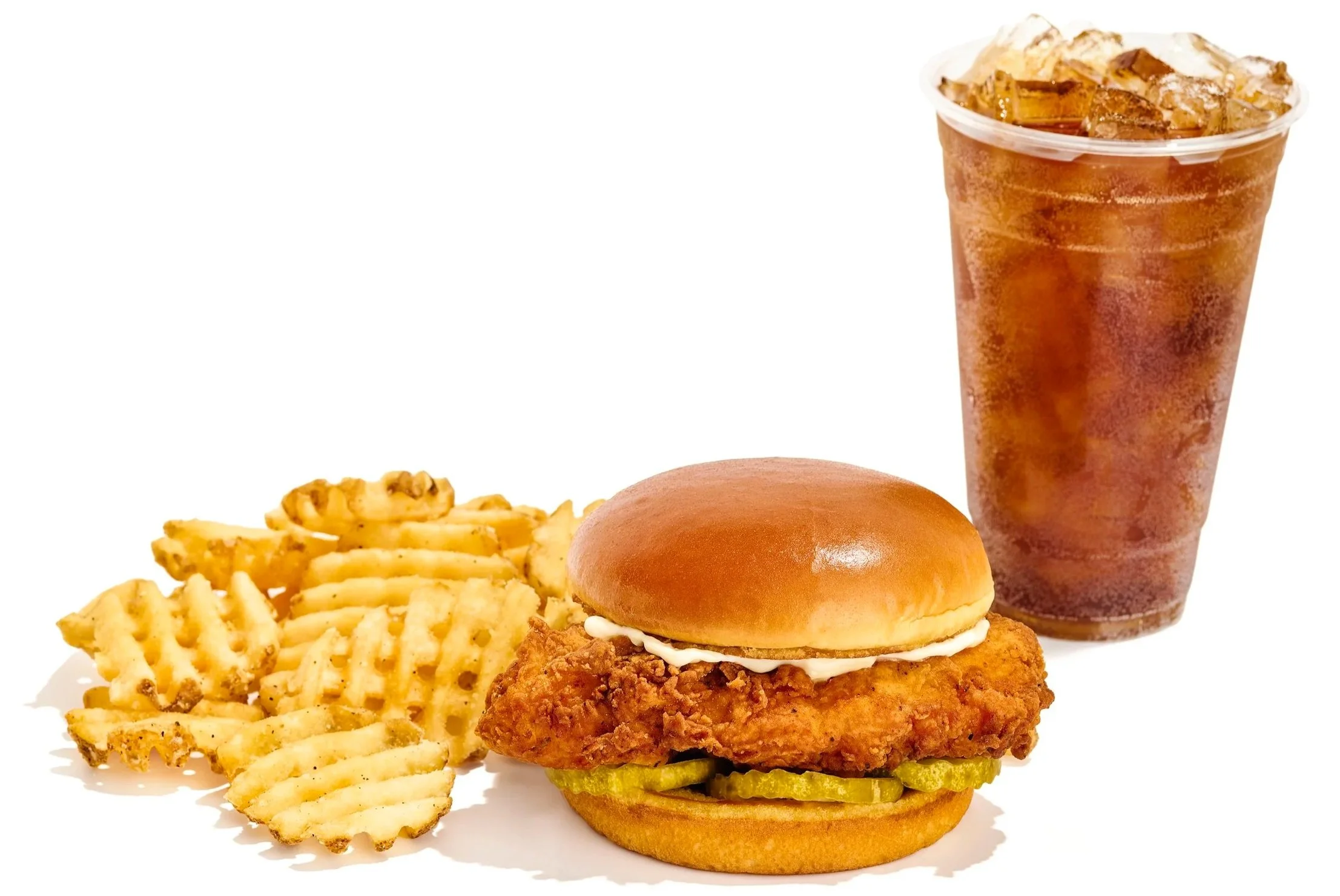 A crispy fried chicken sandwich with pickles, lettuce, and a bun, served with waffle fries and a cold cola in a plastic cup.