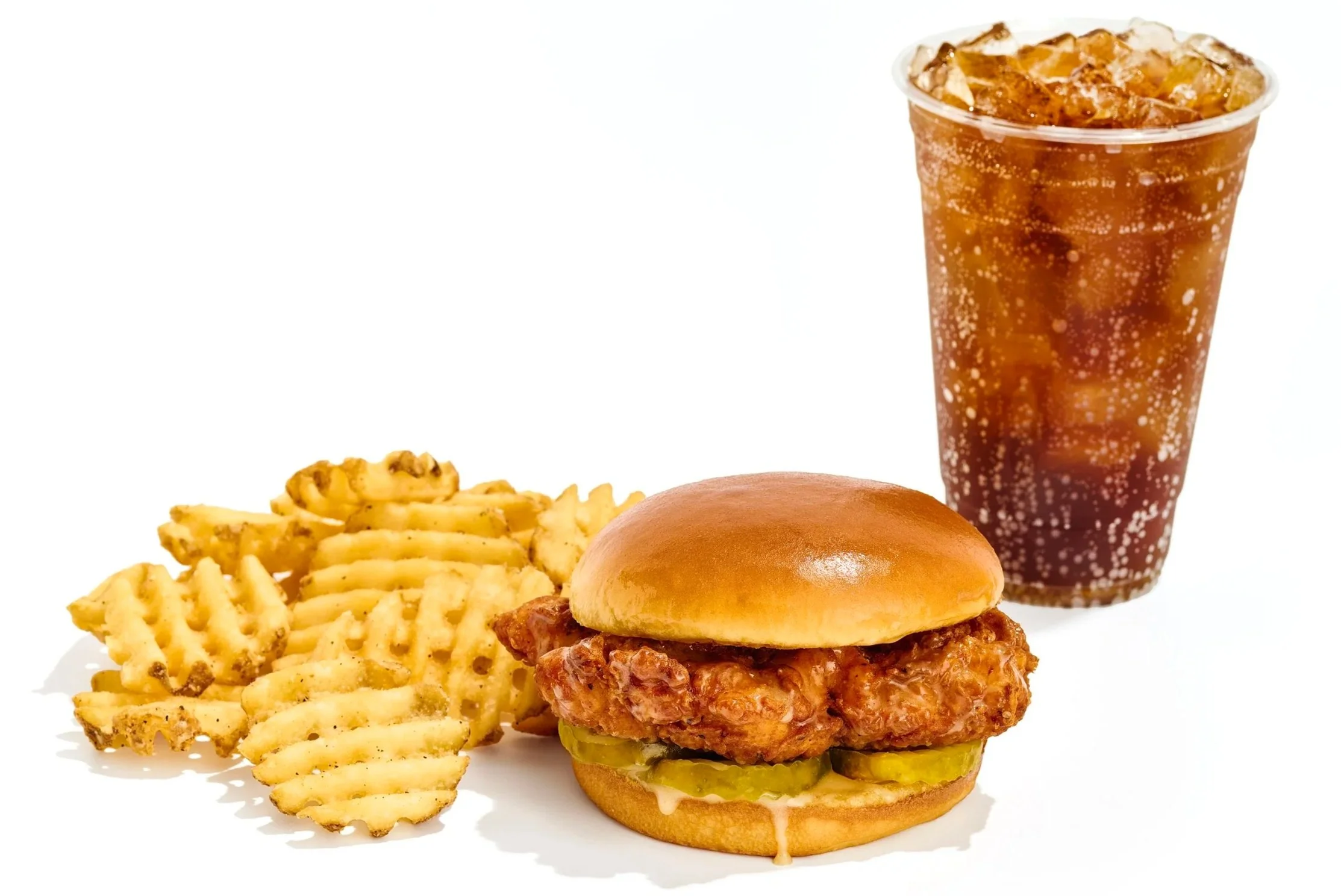 A plate with crispy waffle fries, a chicken sandwich with pickles and sauce, and a soft bun, accompanied by a glass of cola with ice and bubbles.