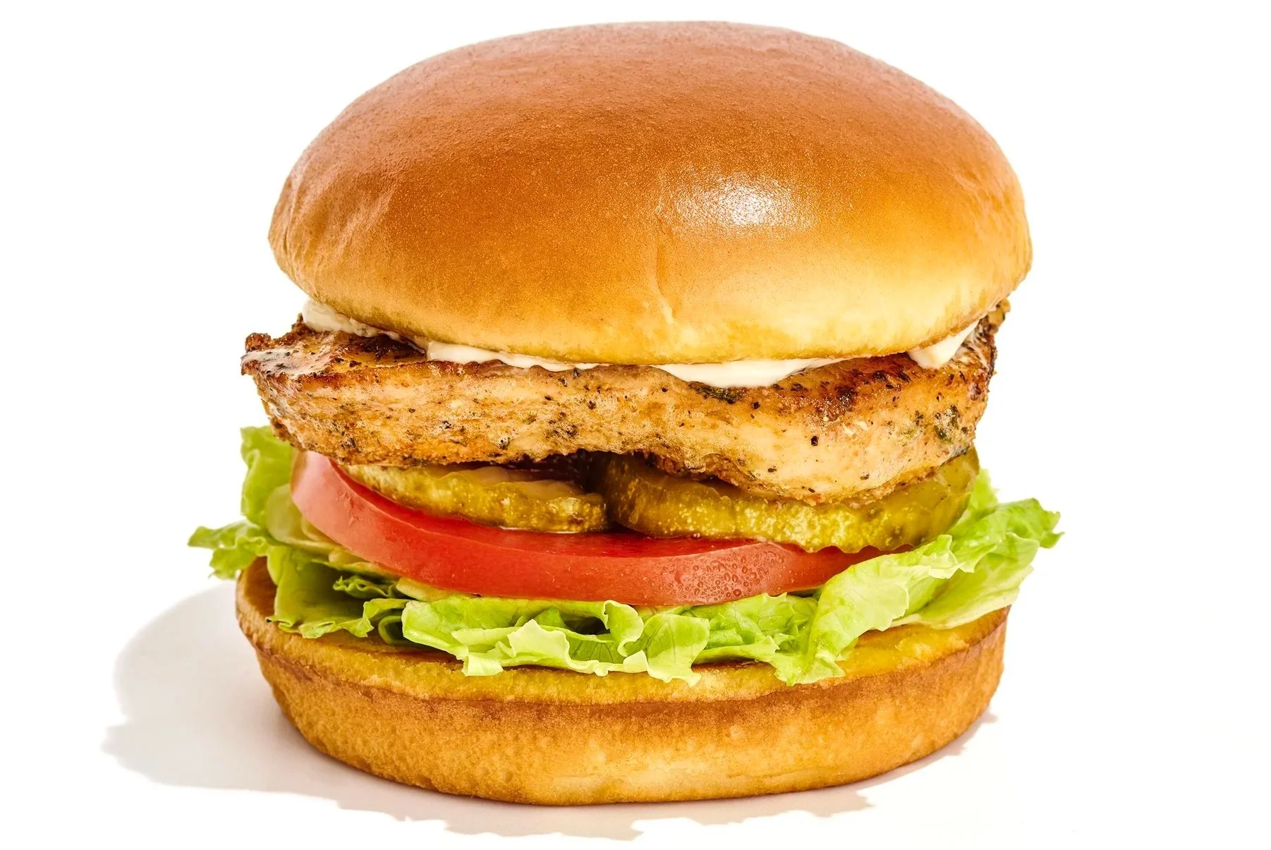 Close-up of a burger with lettuce, tomato, pickles, grilled chicken patty, grilled chicken fillet, mayonnaise, and a bun.